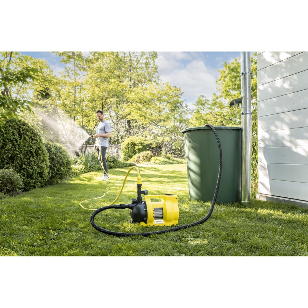 Mann bewässert Garten mit gelber Pumpe neben grüner Regentonne.