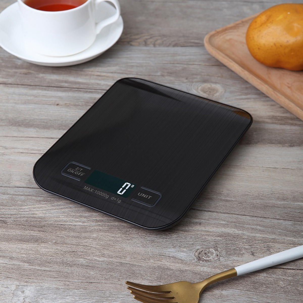 Czarna cyfrowa waga kuchenna na drewnianym stole, obok filiżanka herbaty i jabłko.