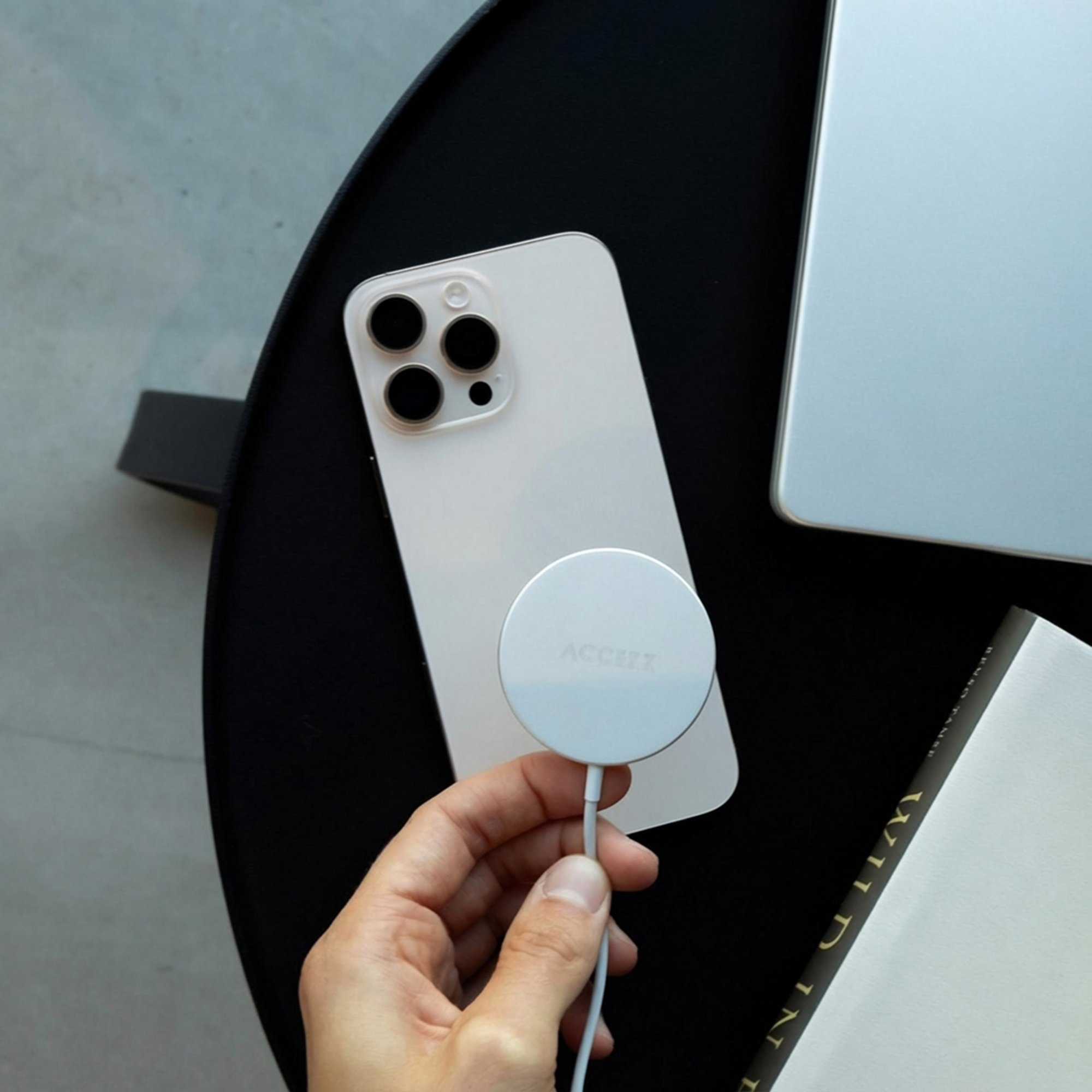Een hand houdt een witte iPhone met een witte lader vast. Een laptop en boek ernaast.