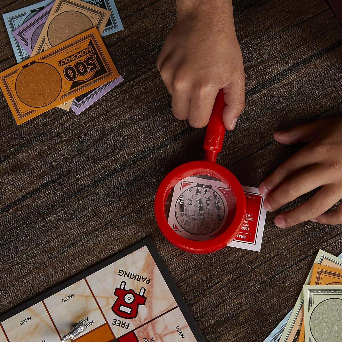 Dłonie używają czerwonej lupy nad kartami i pieniędzmi Monopoly na drewnianym stole.