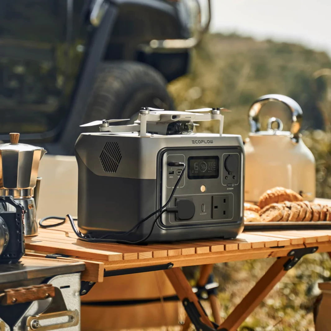 Estación de energía portátil con dron, cafetera, tetera y pan sobre una mesa de madera al aire libre.