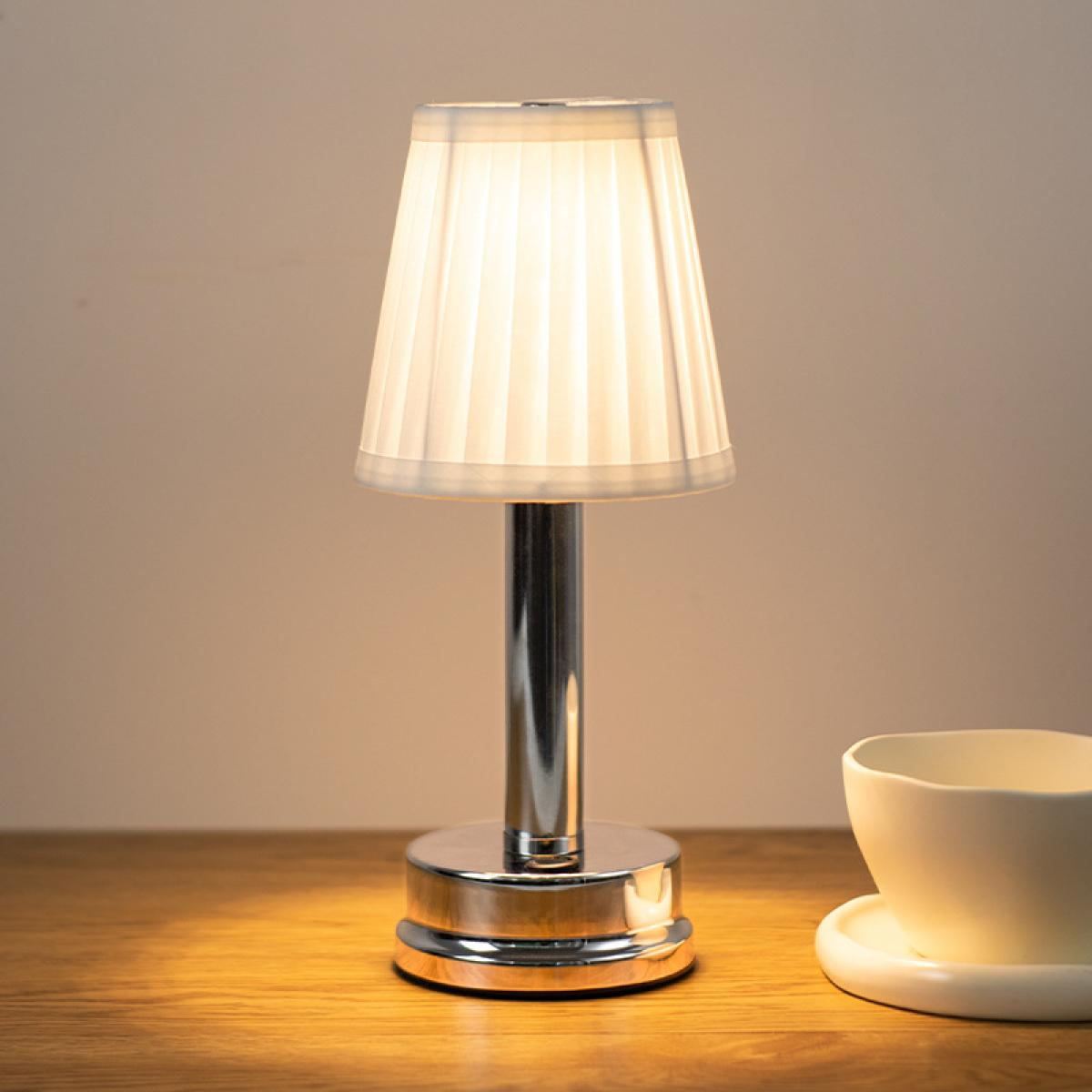 Eine kleine Tischlampe mit Faltenlampenschirm und Metallfuß steht auf einem Holztisch neben einer Tasse.