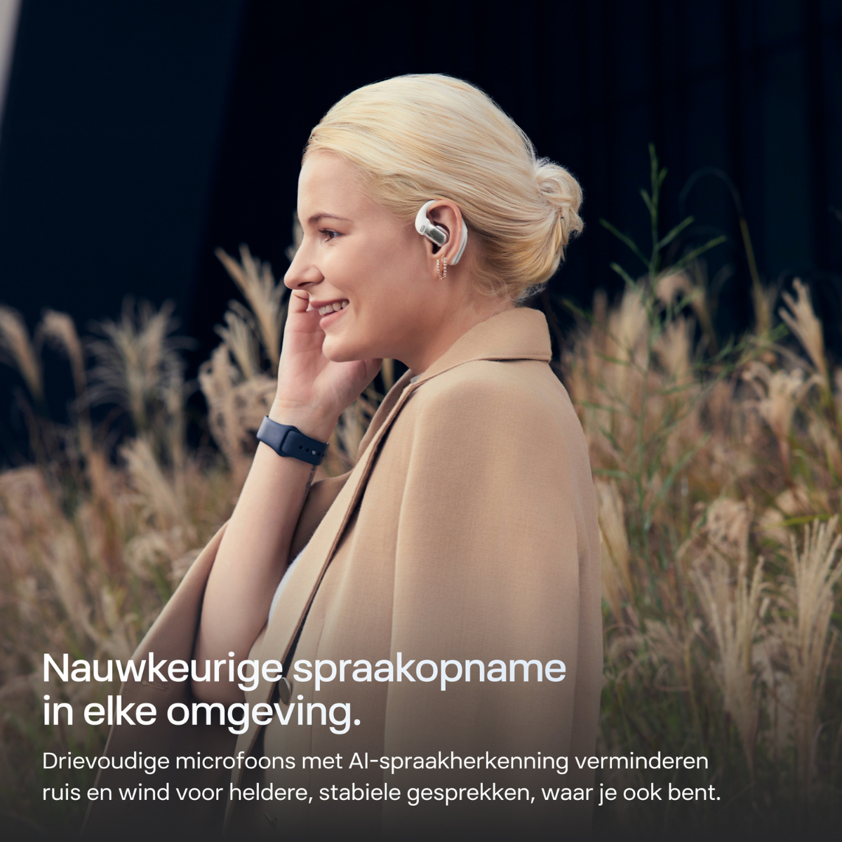 Una donna bionda indossa auricolari e un cappotto beige, sorridendo.