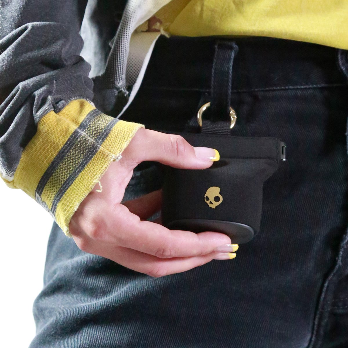 Una mano tiene una custodia nera con logo a teschio, attaccata a jeans neri.