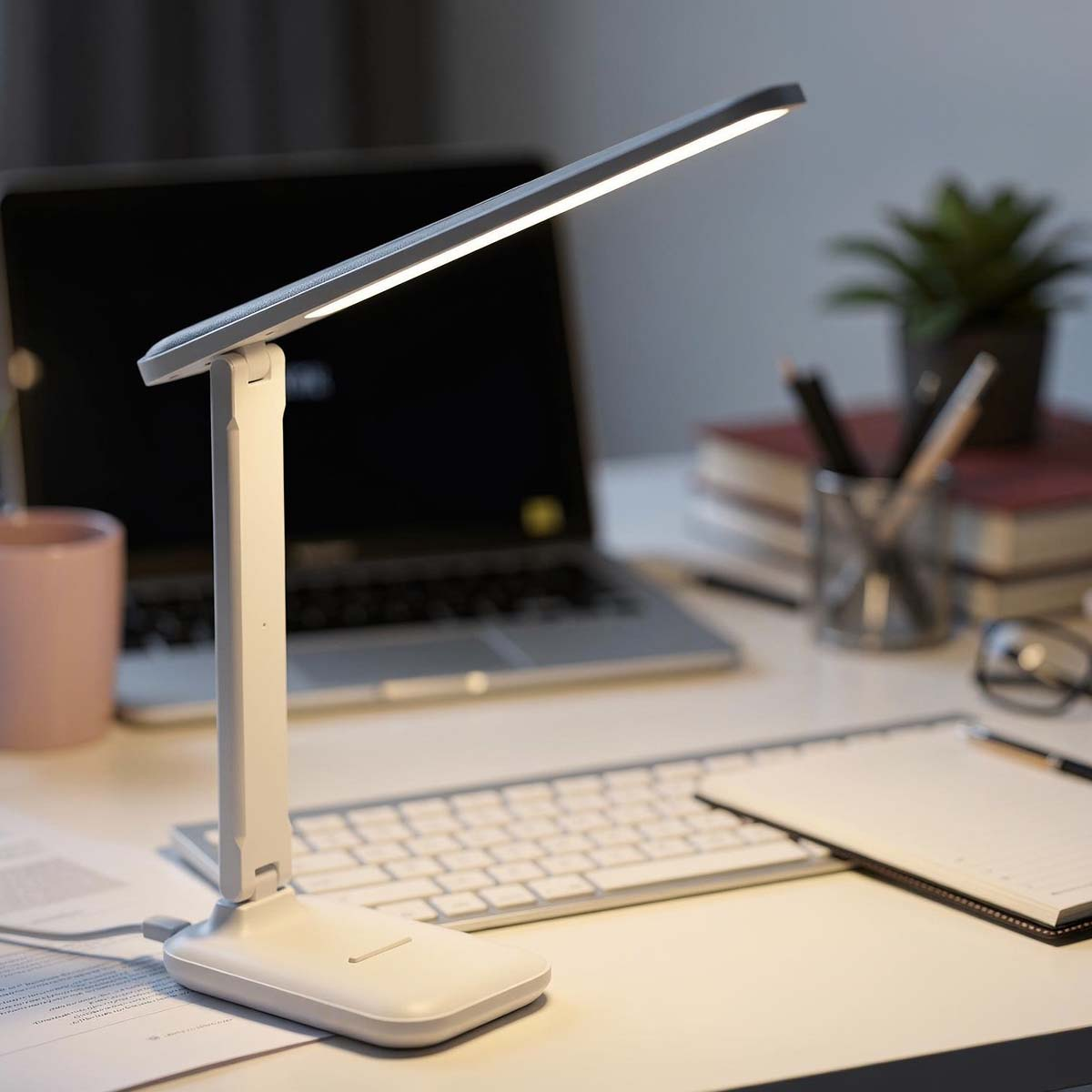 Un escritorio blanco con portátil, teclado, lámpara, cuaderno y planta.