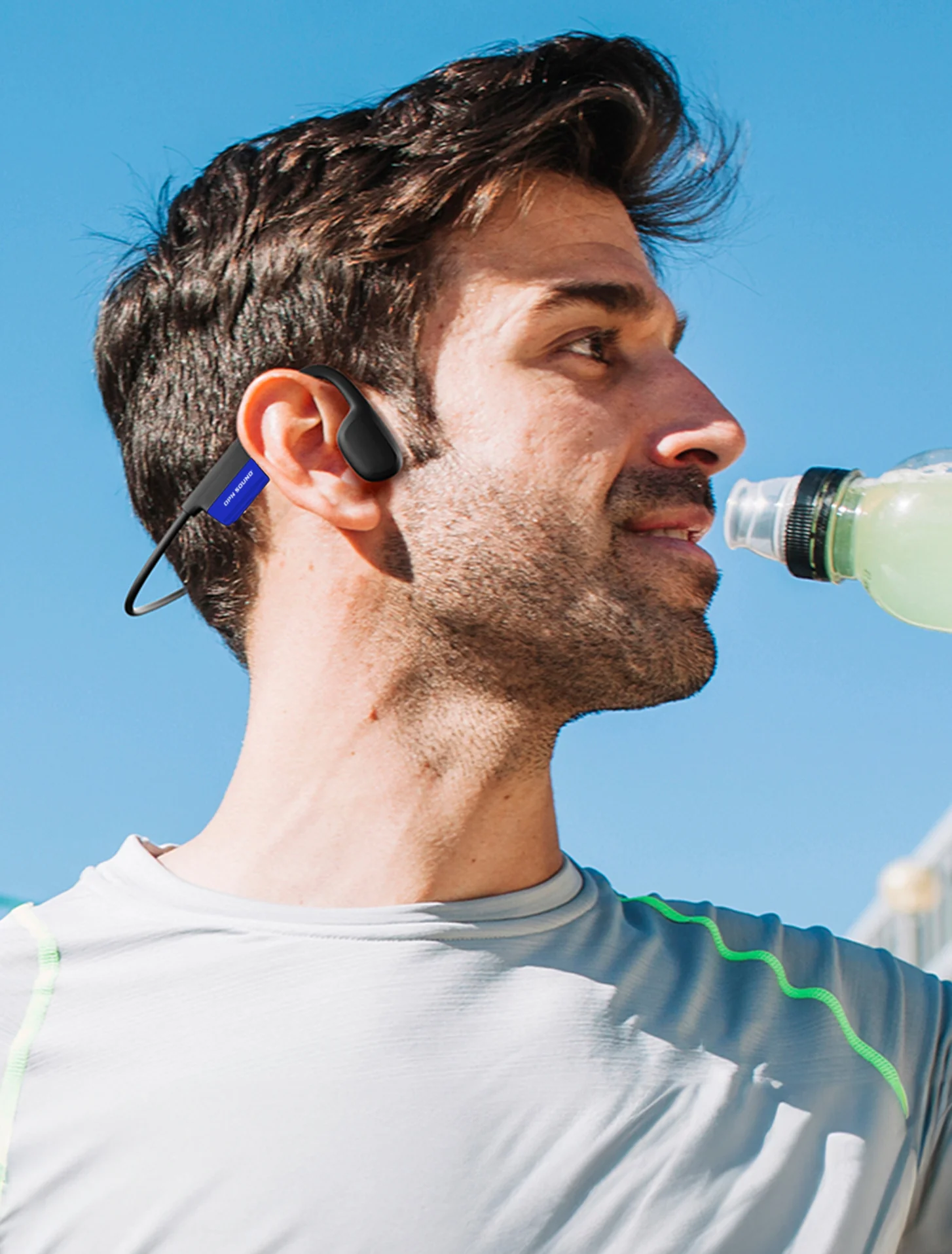 Hombre bebe de una botella verde, con auriculares y camiseta gris.