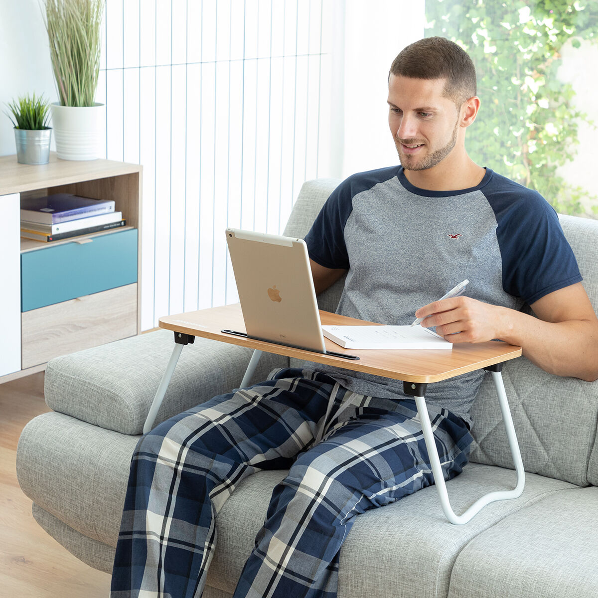 Mann sitzt auf Sofa mit Laptop-Tisch, arbeitet am Tablet.