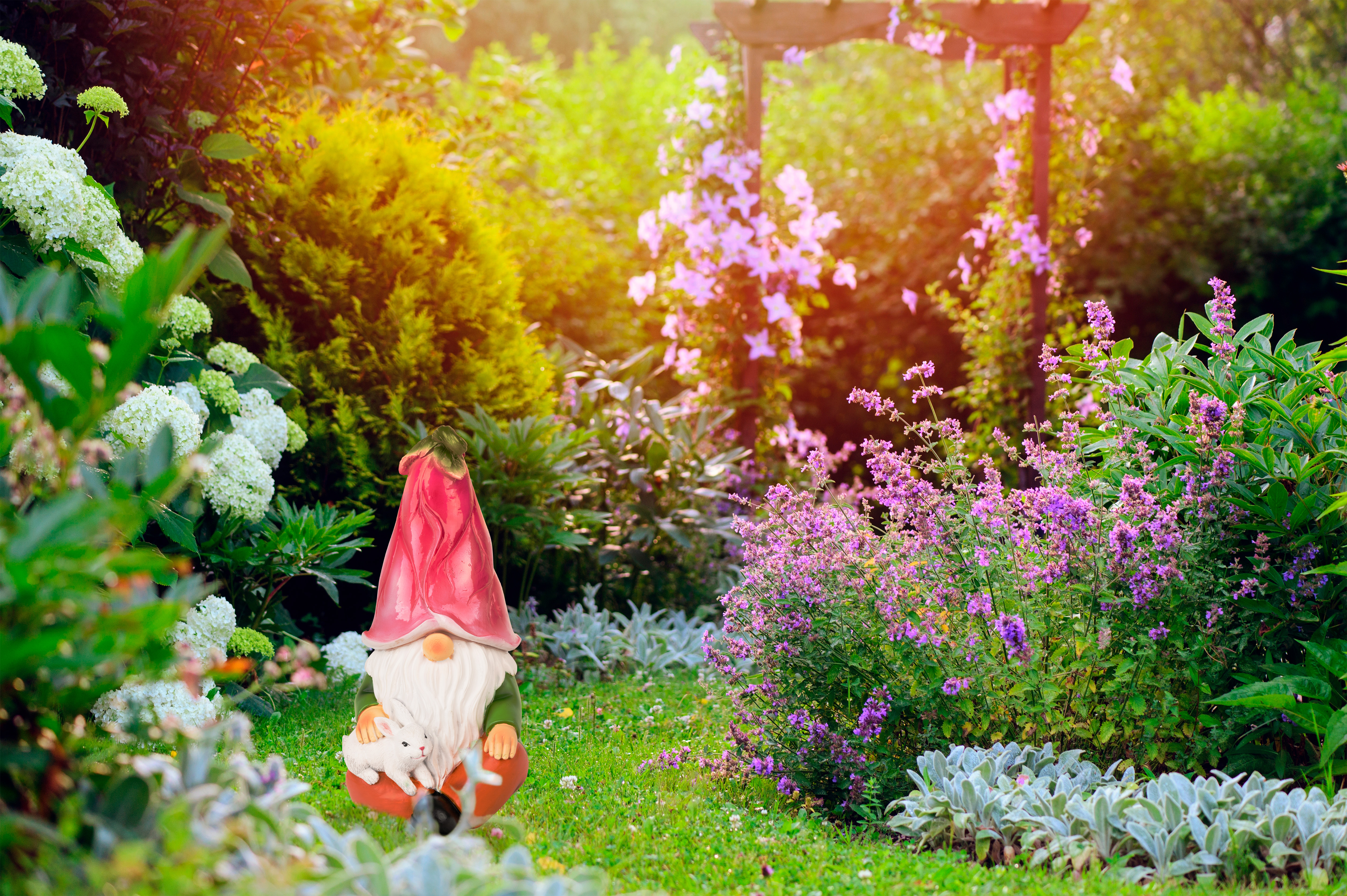 Ein Gartenzwerg mit einem Hasen sitzt in einem üppigen Garten mit lila und weißen Blumen.