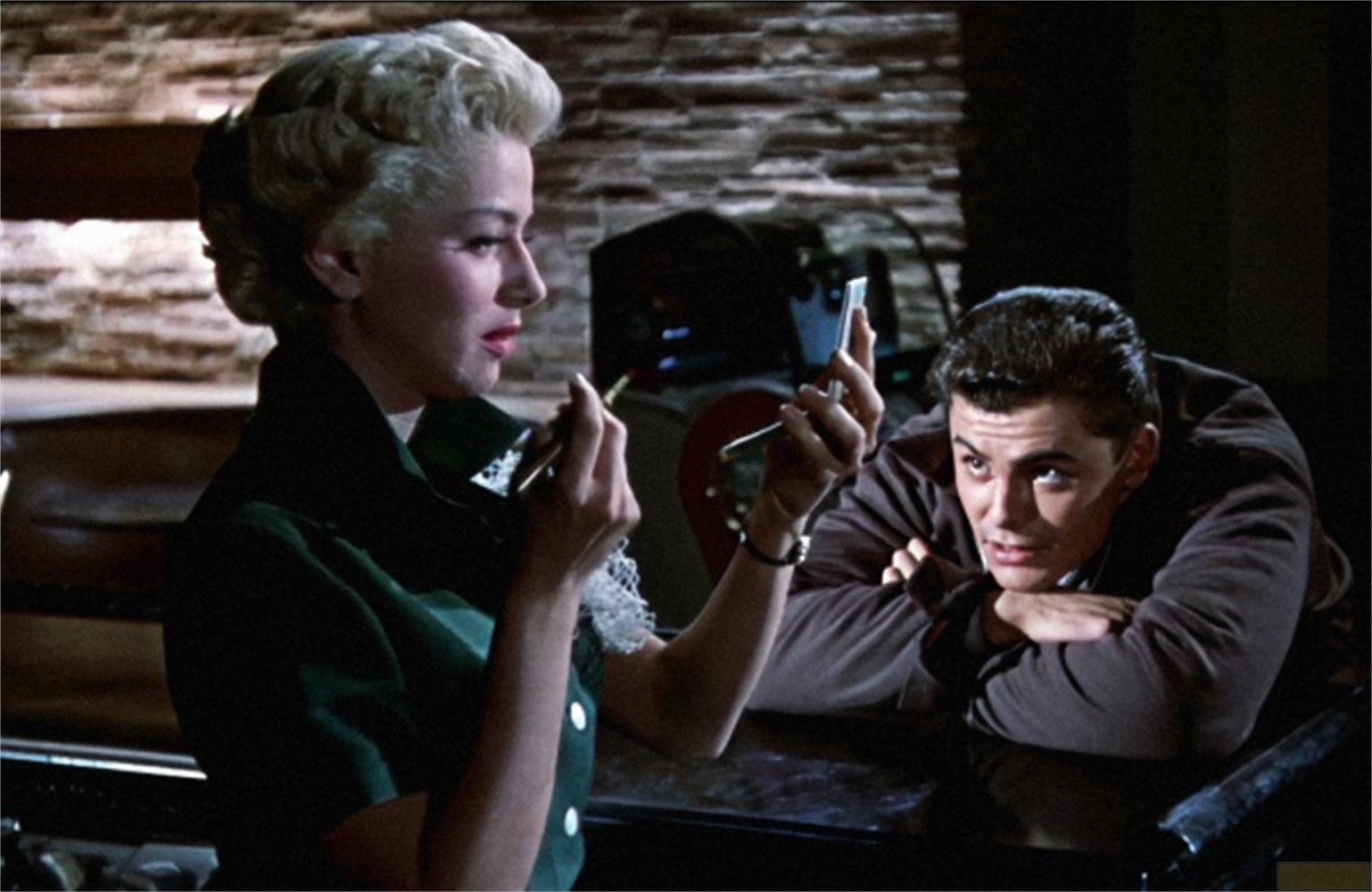 Una donna bionda si trucca con il rossetto mentre un uomo la osserva.
