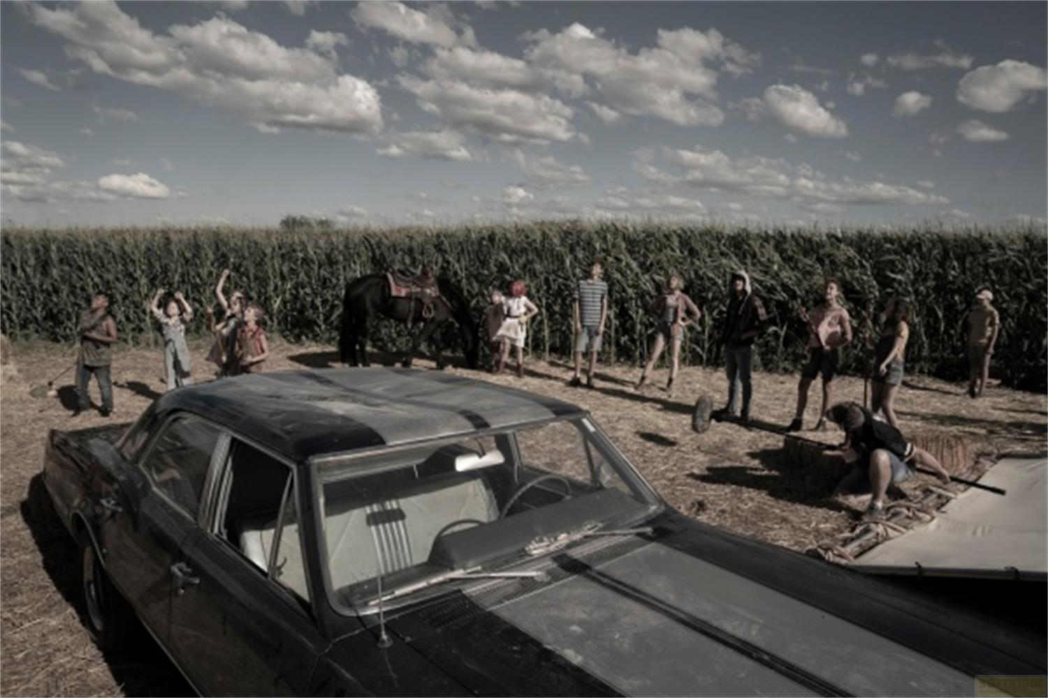 Un'auto d'epoca davanti a un campo di mais, con persone e un cavallo sullo sfondo.