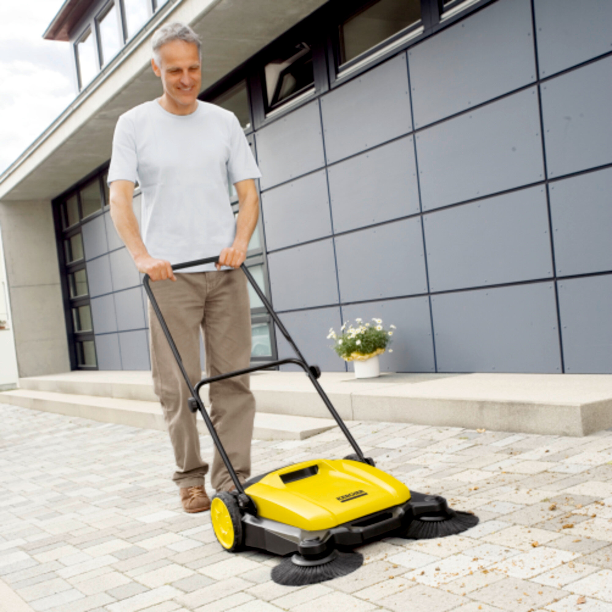 Mann schiebt gelbe Kehrmaschine auf gepflastertem Weg vor modernem Haus.