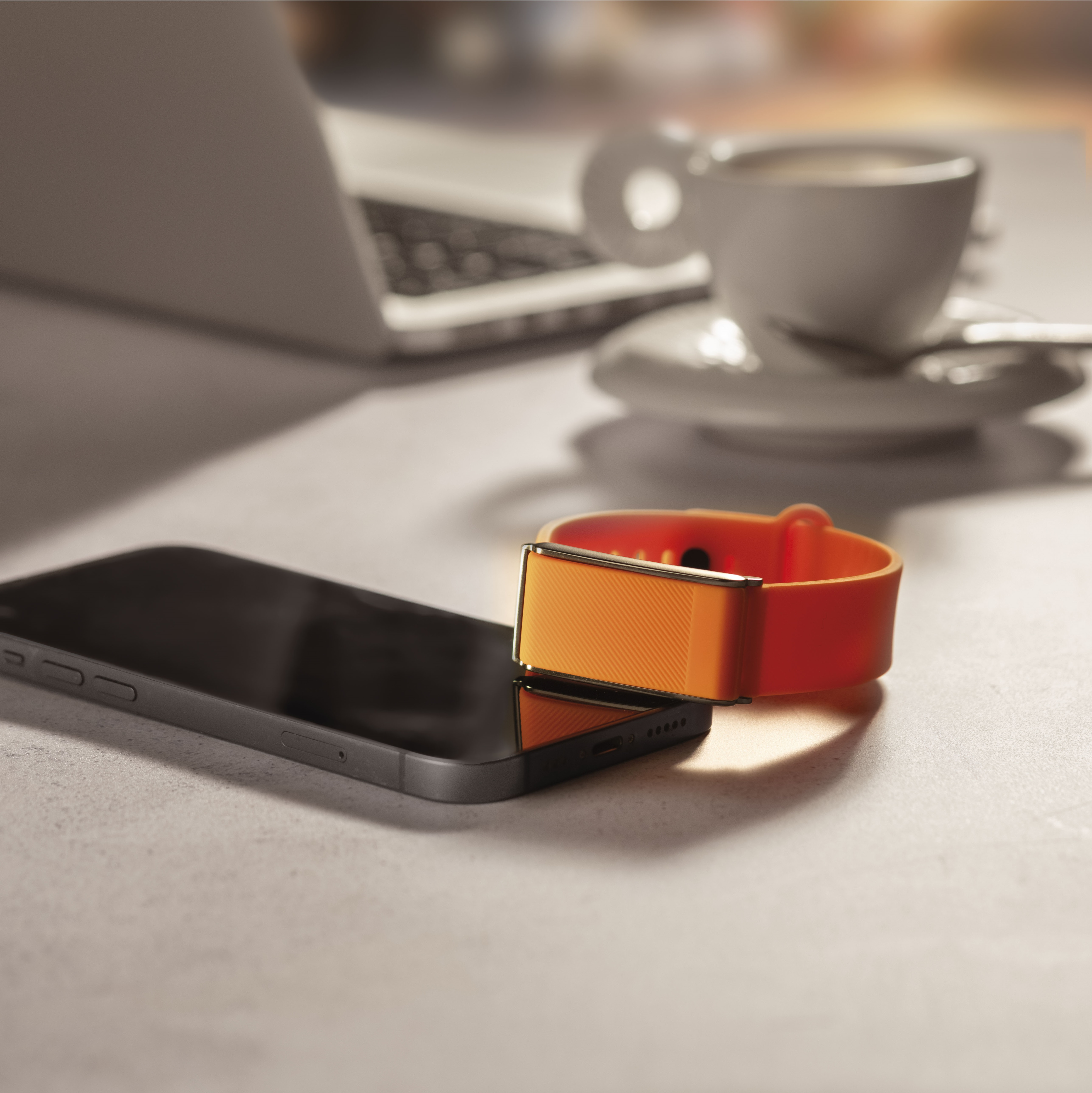 Una pulsera de actividad naranja descansa sobre un smartphone, con café y portátil cerca.