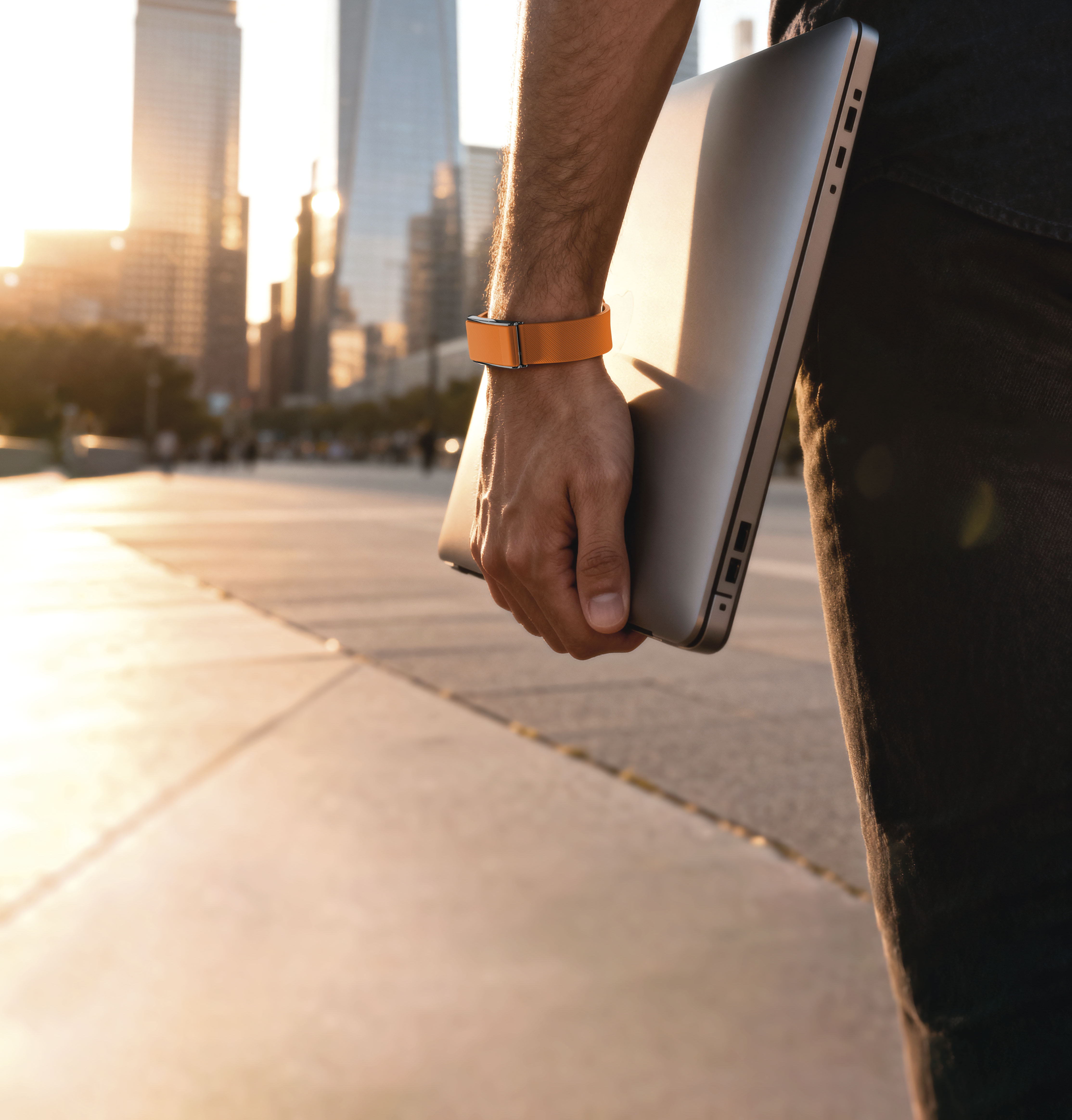 Hombre sostiene portátil con pulsera naranja, horizonte urbano al atardecer.