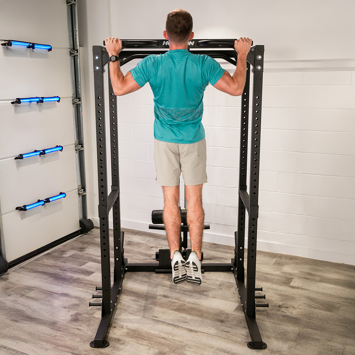 Hombre haciendo dominadas en un power rack negro en un gimnasio en casa con luces azules en la pared.