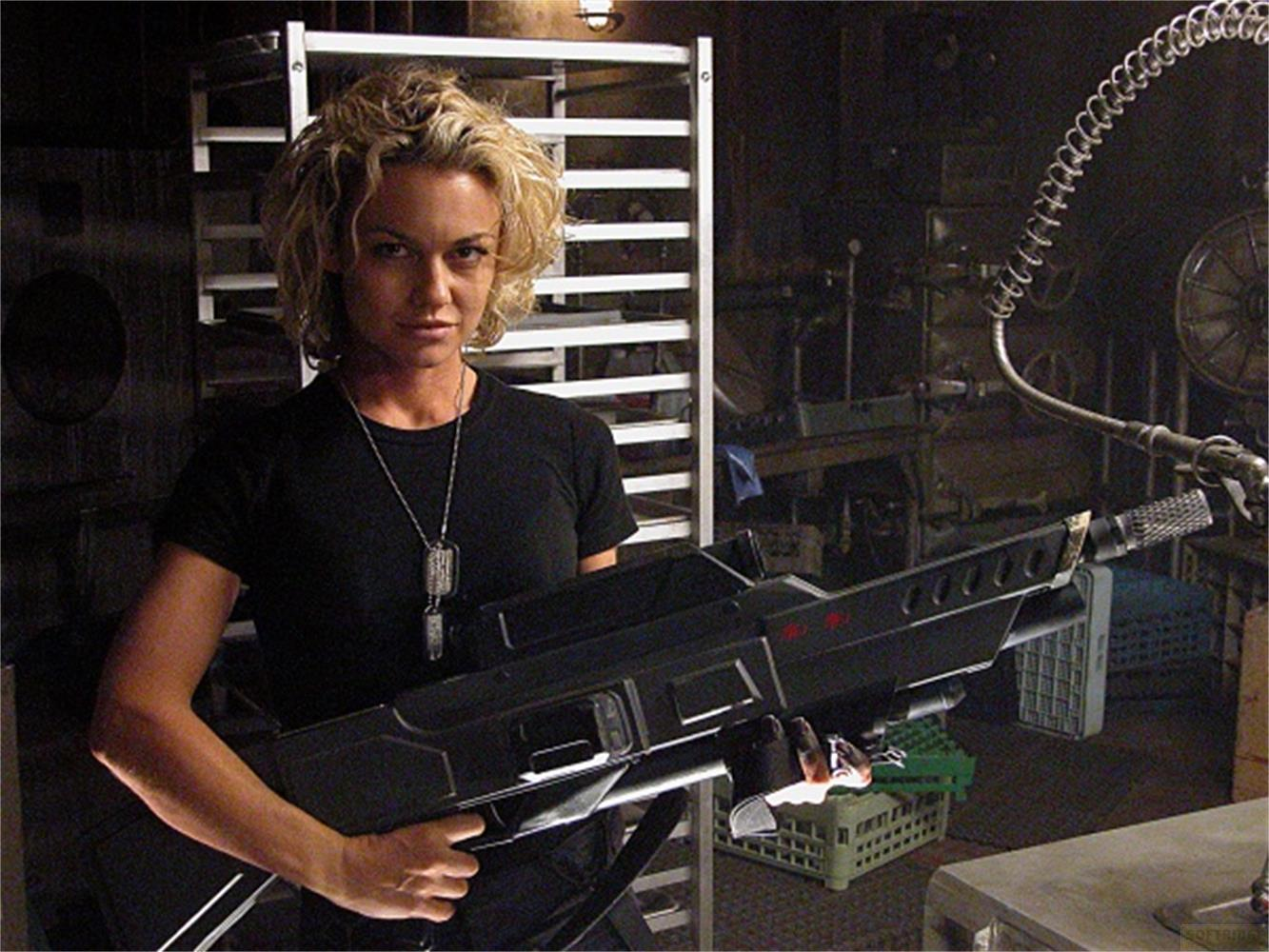 Donna con capelli biondi ricci in maglia nera tiene fucile nero futuristico. Sfondo scuro e industriale.
