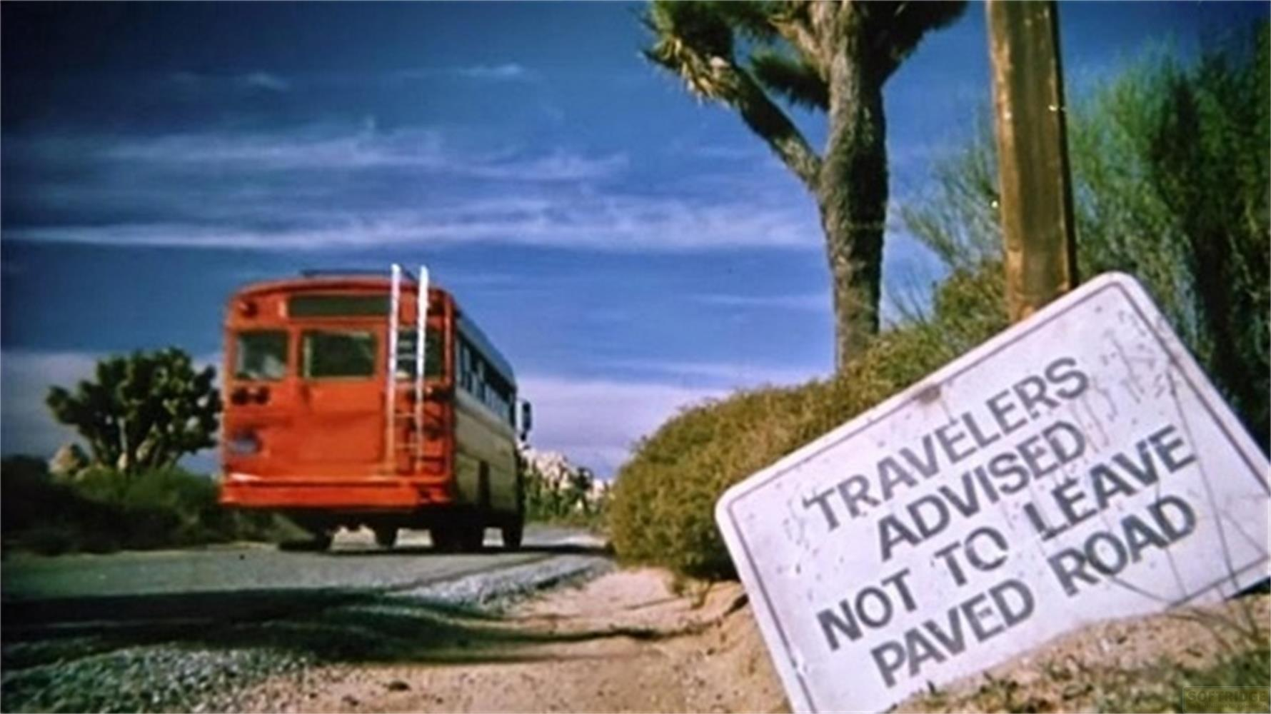 Un autobus rosso percorre una strada desertica, un cartello avverte di non lasciare la strada.