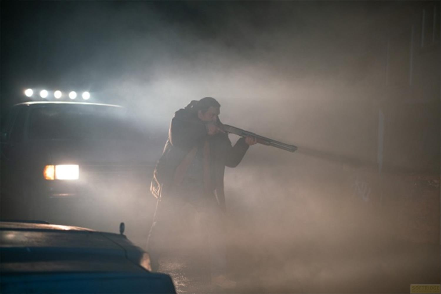 Uomo con fucile nella nebbia accanto a un'auto con luci.