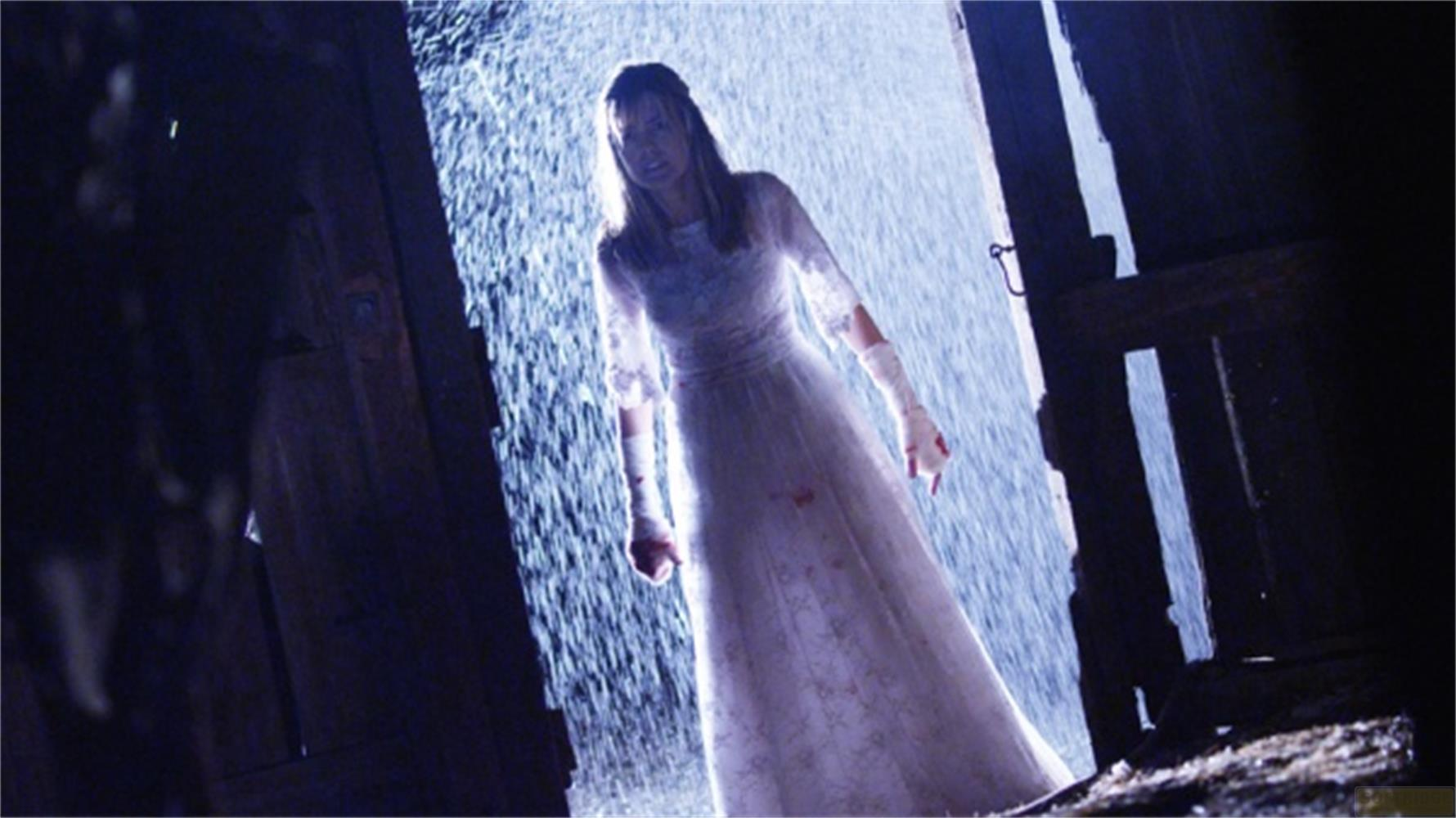 Una donna in abito bianco sta sotto la pioggia davanti a un ingresso scuro.