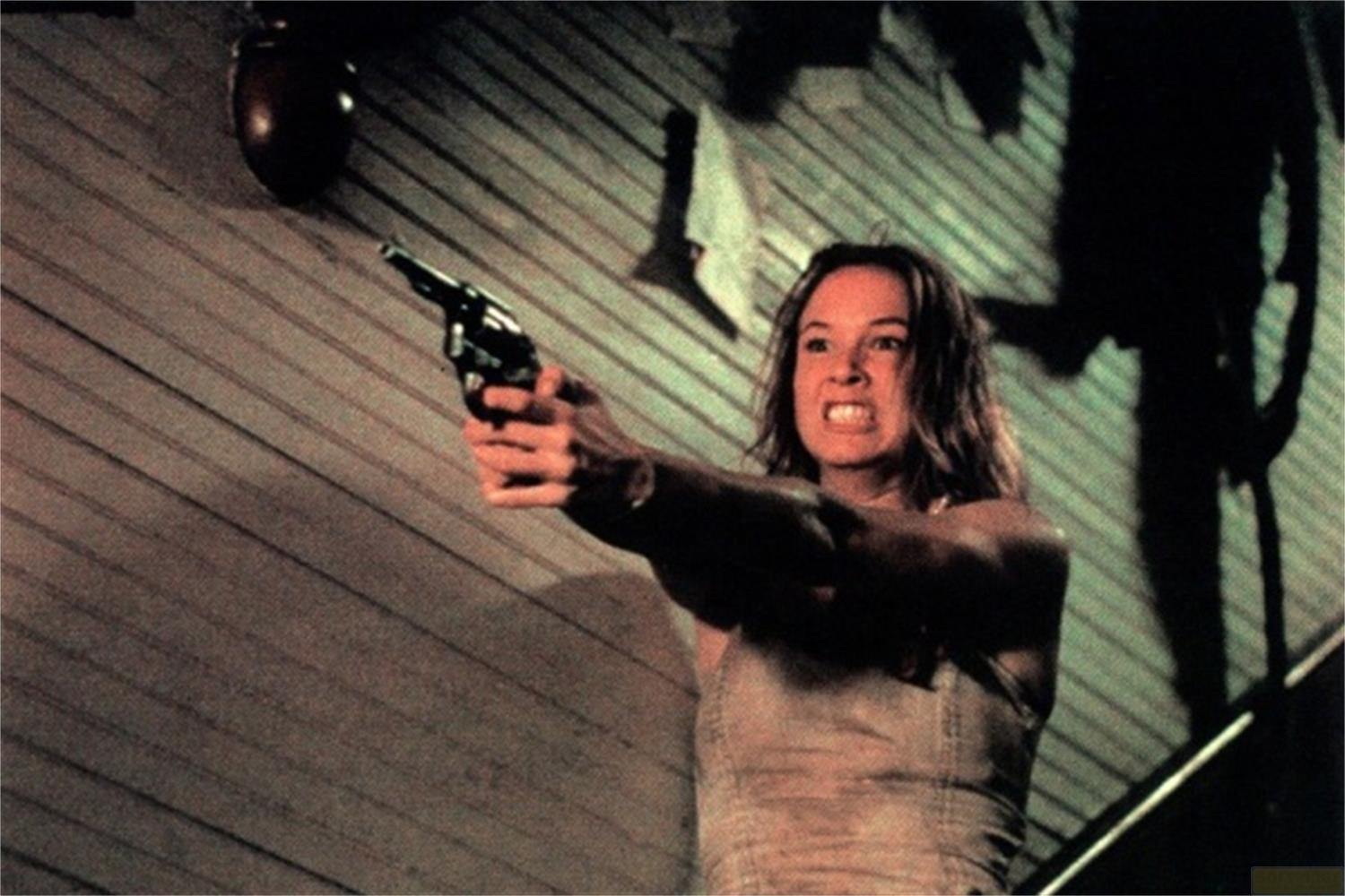 Una donna con faccia arrabbiata tiene una pistola. Muro di legno sullo sfondo.