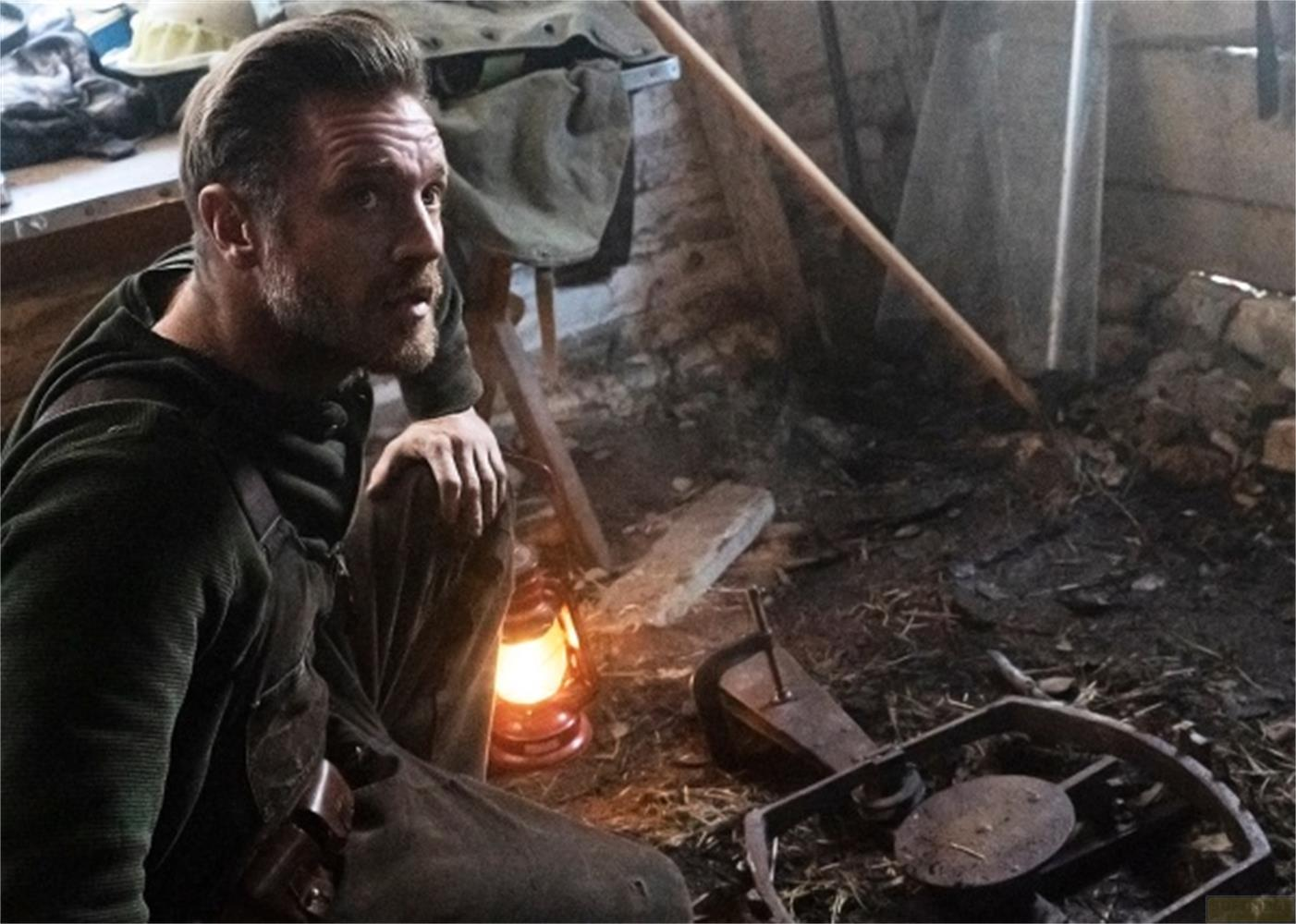 Un uomo si inginocchia accanto a una lanterna accesa in una stanza rustica.