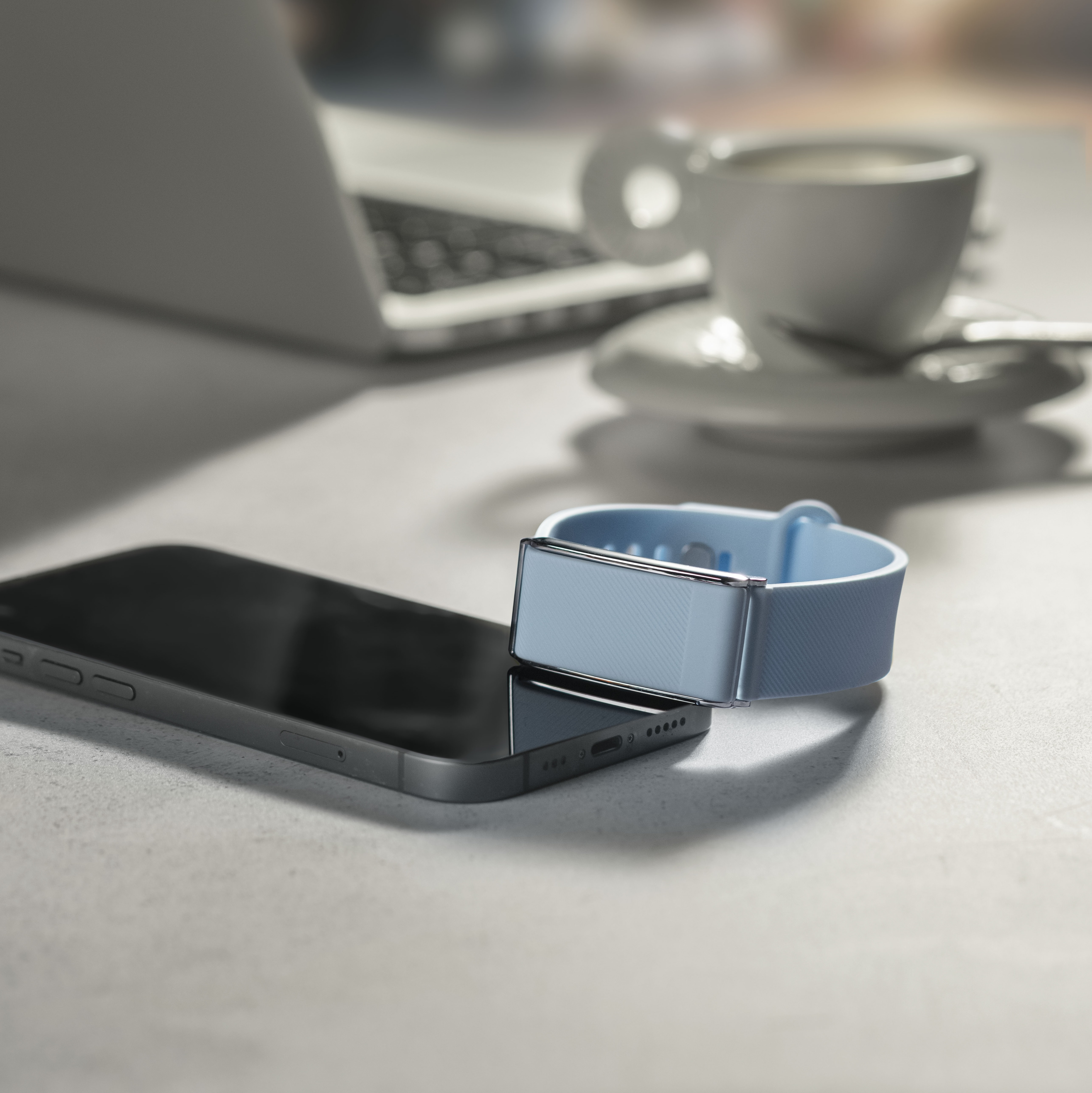 Una pulsera de actividad azul descansa sobre un smartphone, con taza de café y portátil cerca.