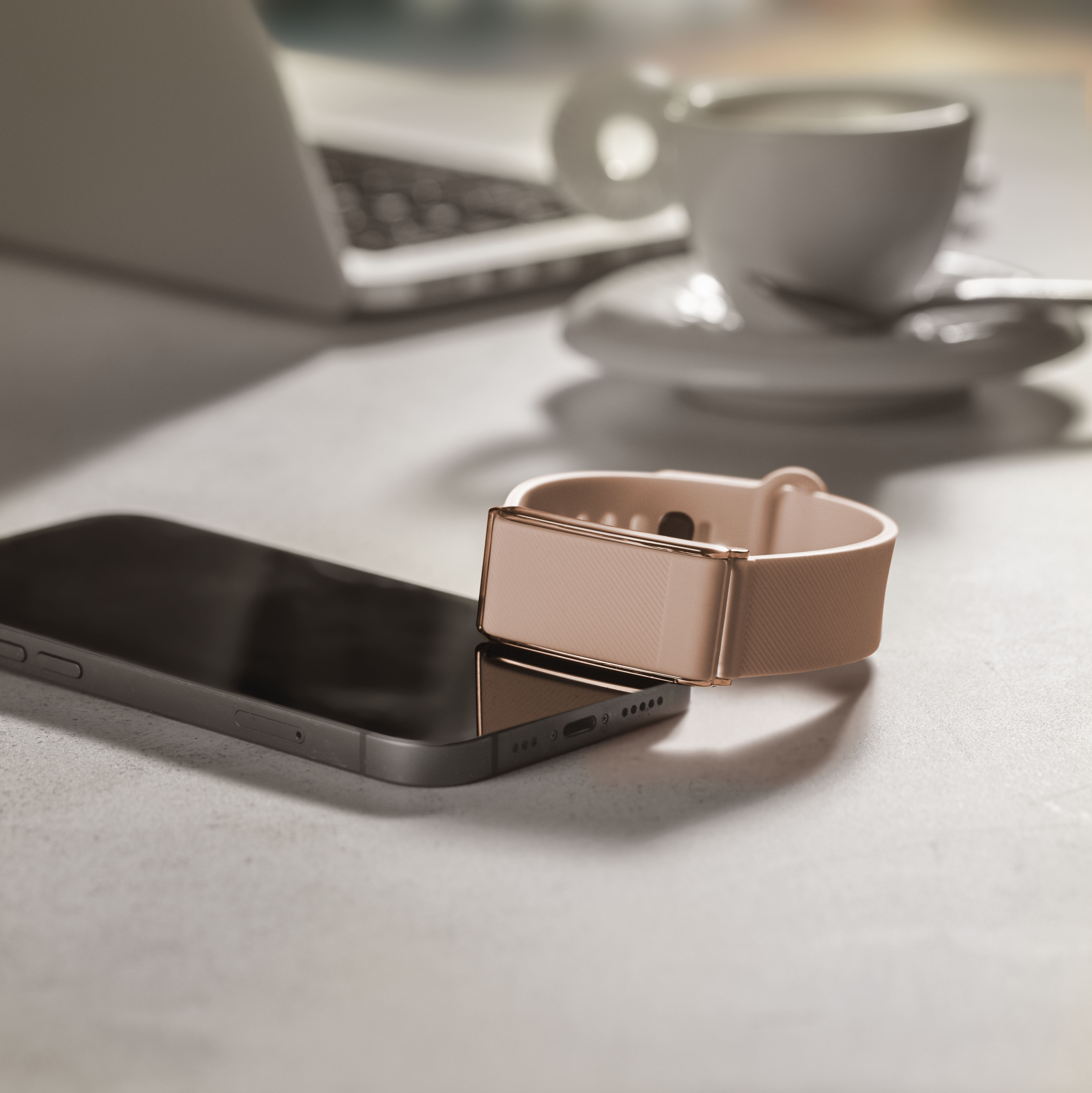 Una pulsera de actividad en oro rosa descansa sobre un smartphone, con portátil y café.