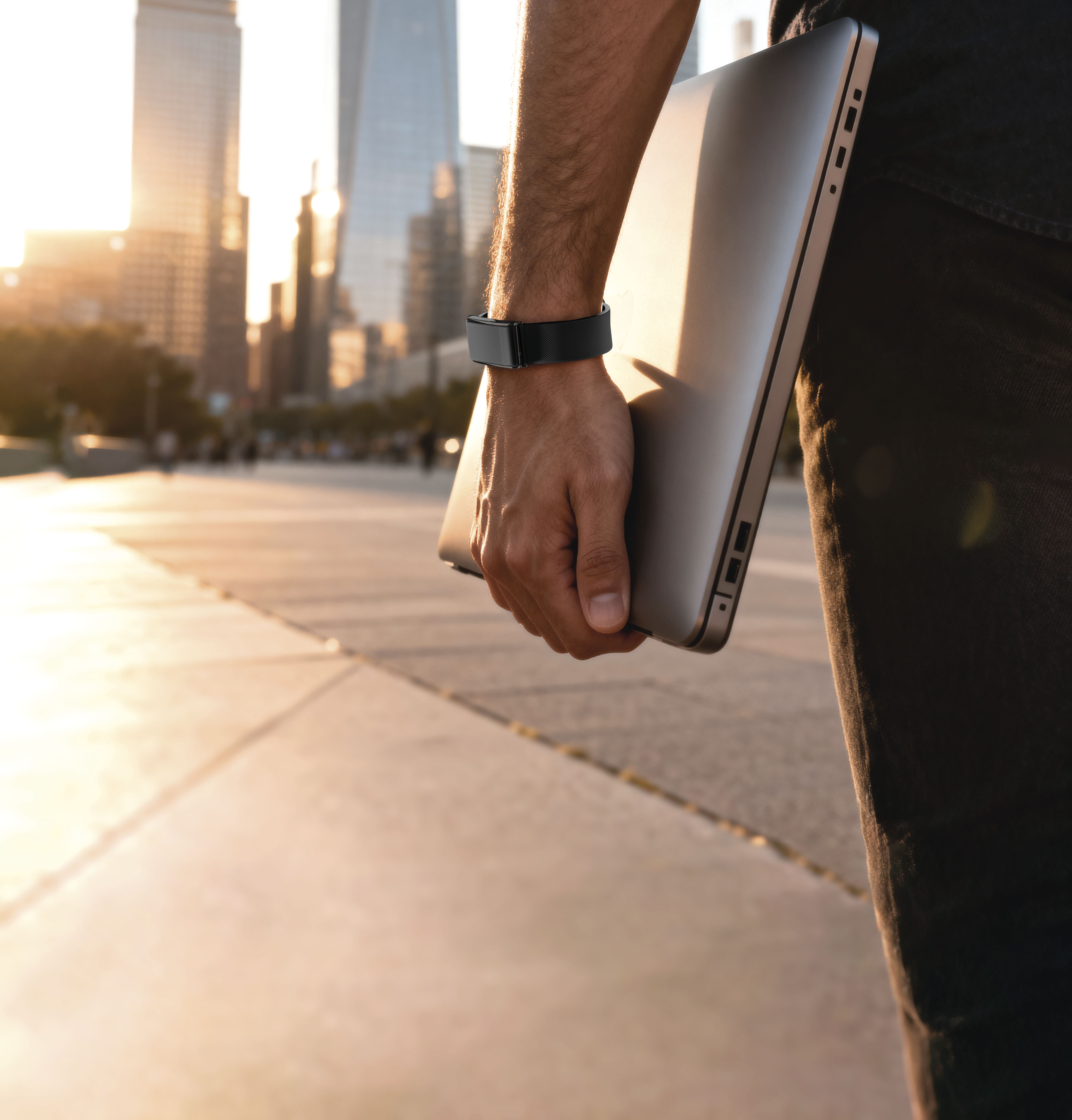 Hombre con portátil, lleva smartwatch, ante skyline al atardecer.