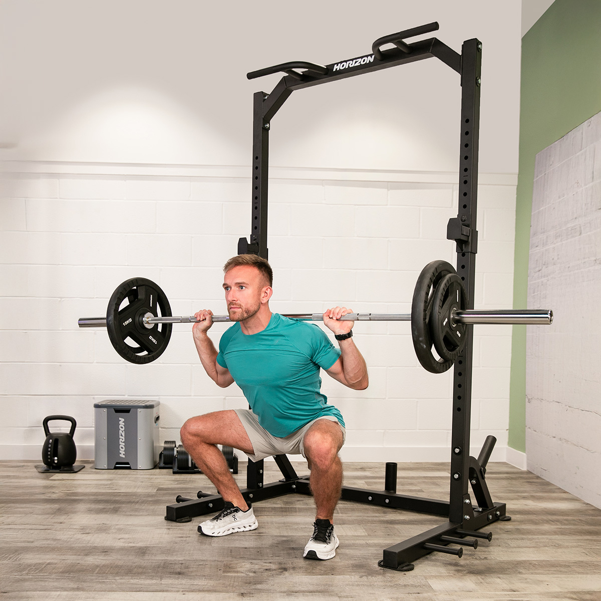 Hombre hace sentadillas con barra bajo un rack en un gimnasio. Pesa rusa y caja cerca.