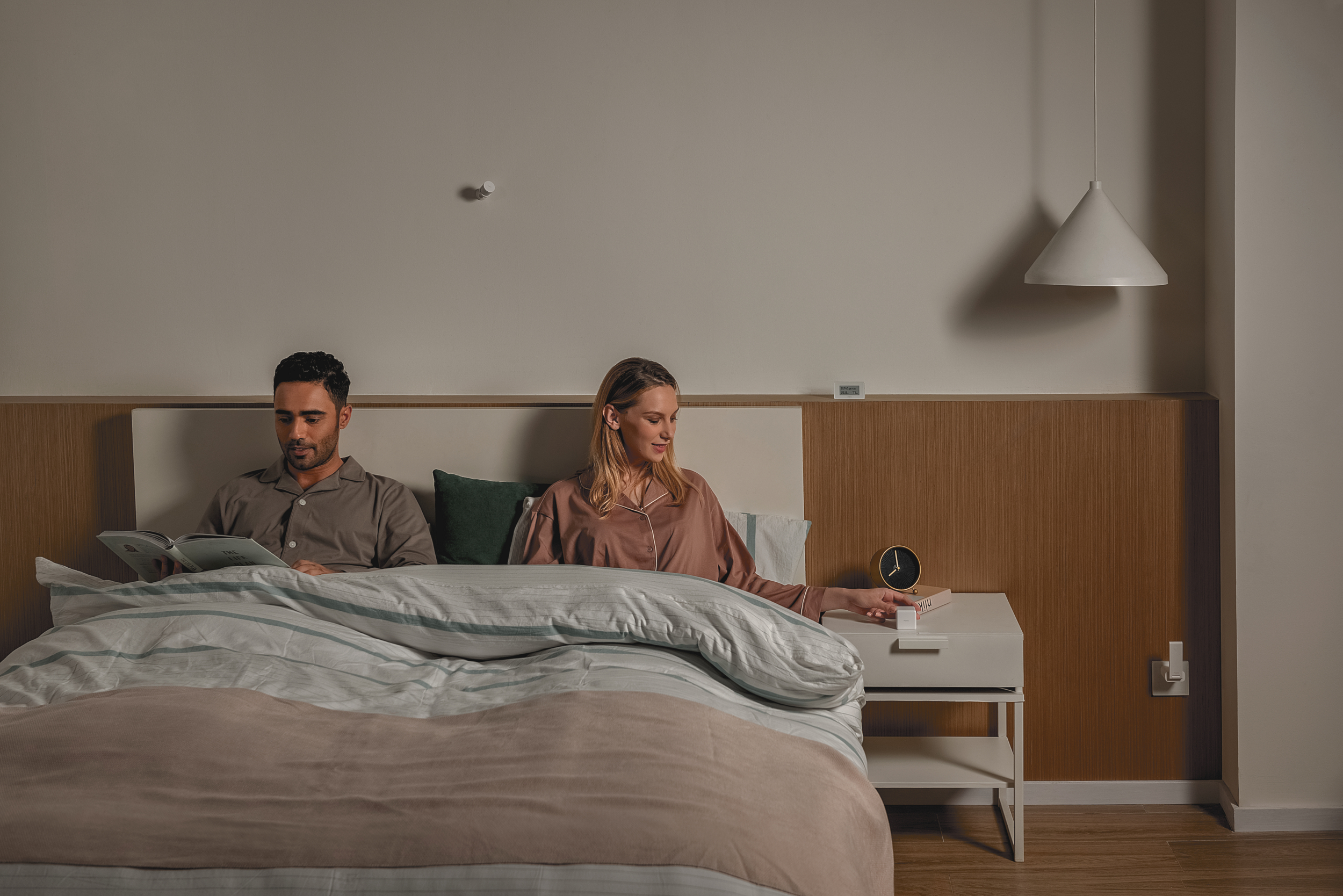 Un hombre lee en la cama, una mujer alcanza un interruptor en una mesita de noche.