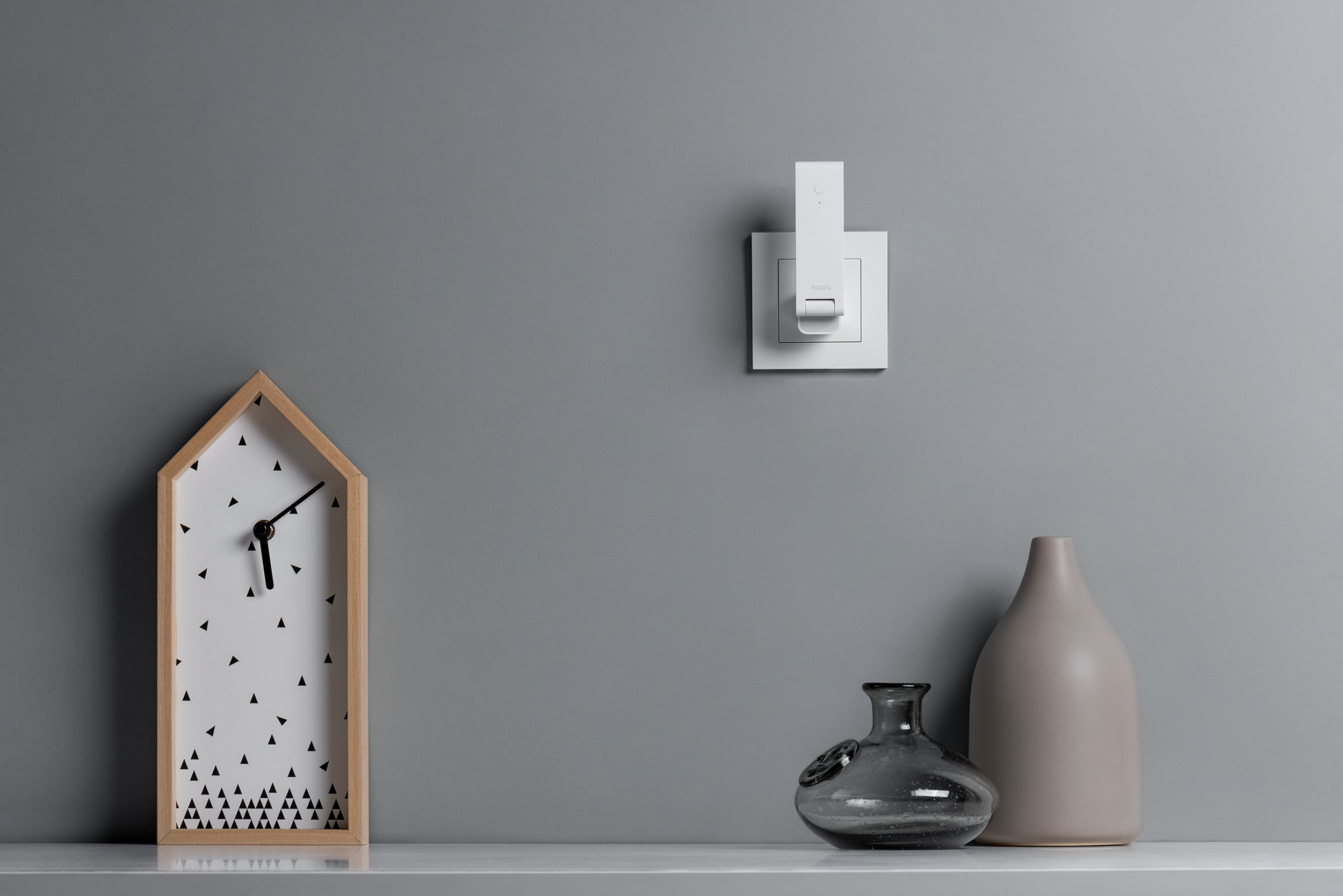 Un reloj con forma de casa, un interruptor blanco y dos jarrones en un estante.