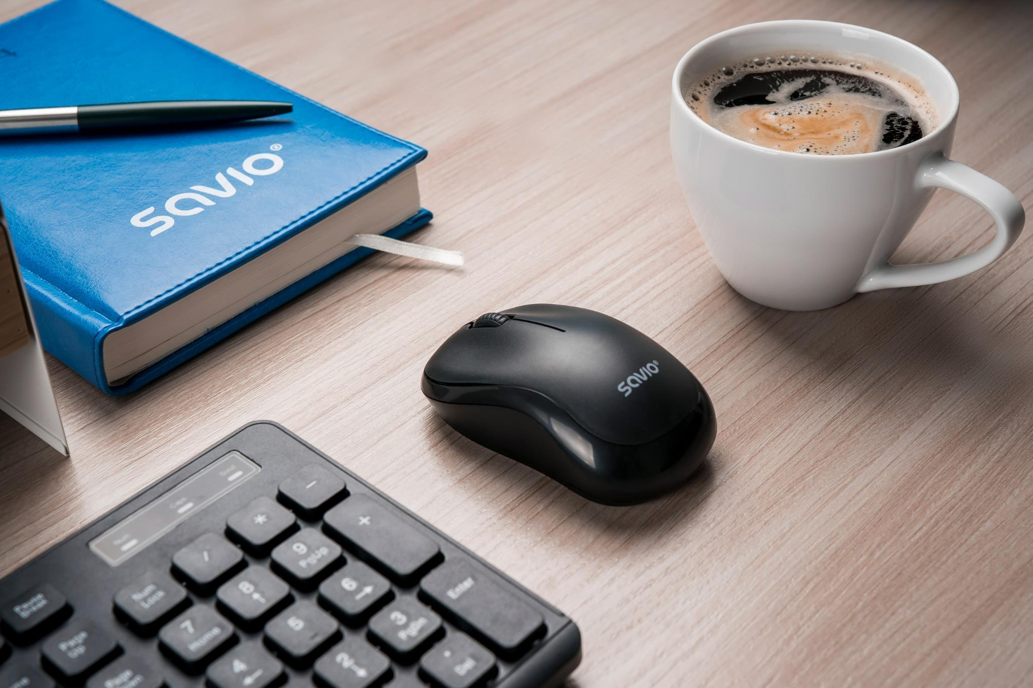 Niebieski notatnik, czarna mysz, filiżanka kawy i klawiatura na drewnianym biurku.