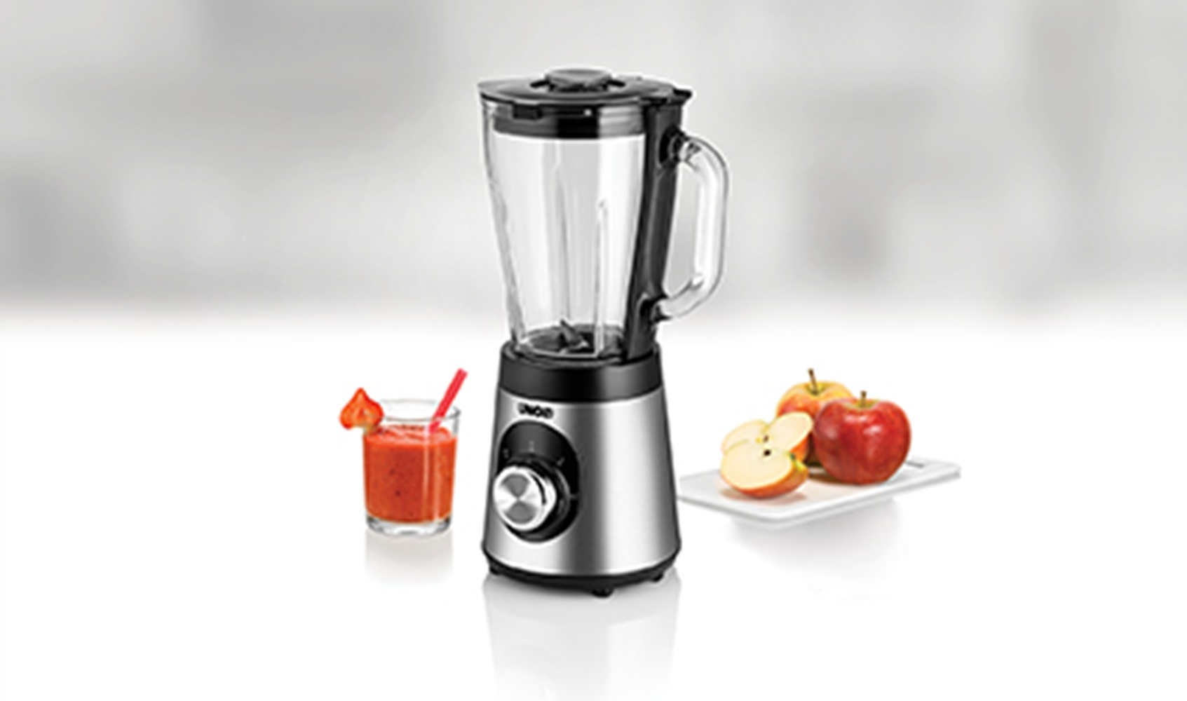 Ein silberner Mixer mit Glasbehälter neben einem Glas Smoothie und Äpfeln.
