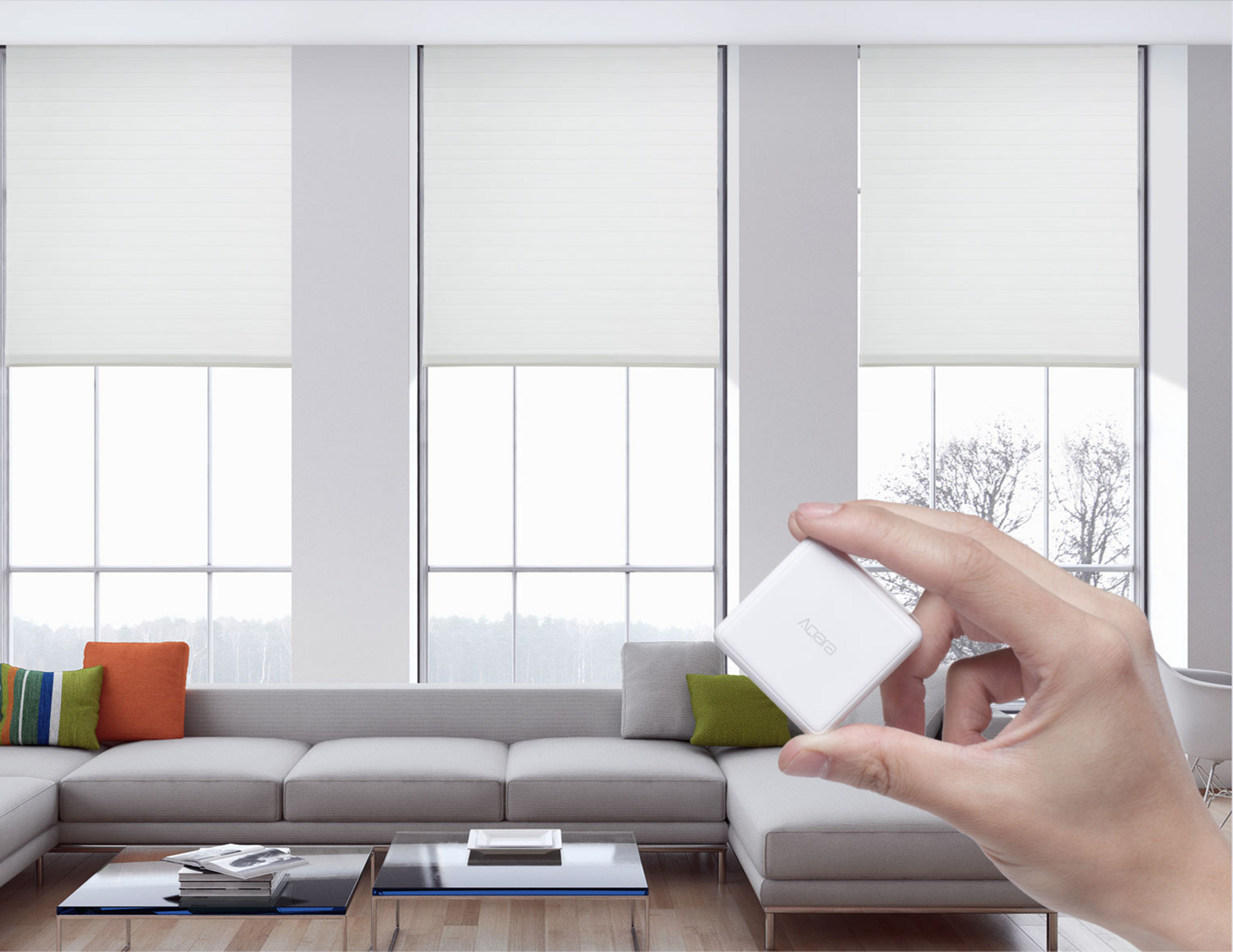 Ręka trzyma białe urządzenie Aqara w salonie z sofą i dużymi oknami.