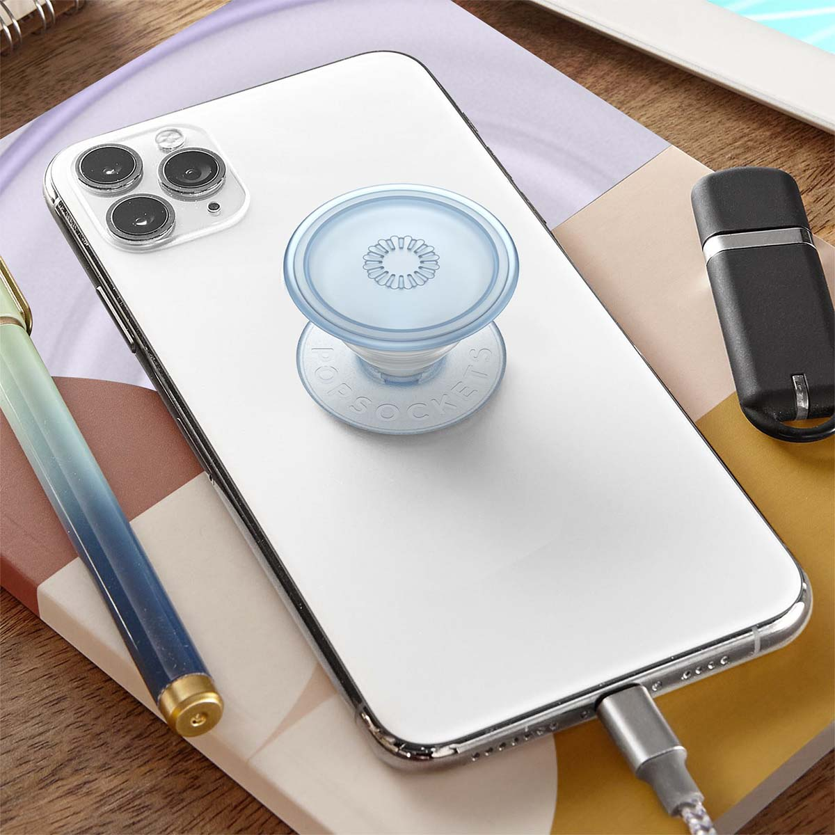 Un smartphone blanco con un Pop-Socket azul, cable de carga y bolígrafo sobre una superficie colorida.