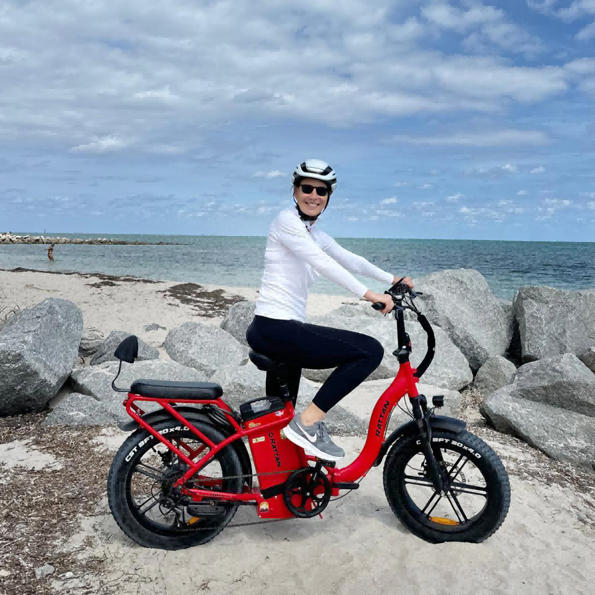 Kobieta na czerwonym rowerze elektrycznym na plaży z kamieniami i morzem w tle.