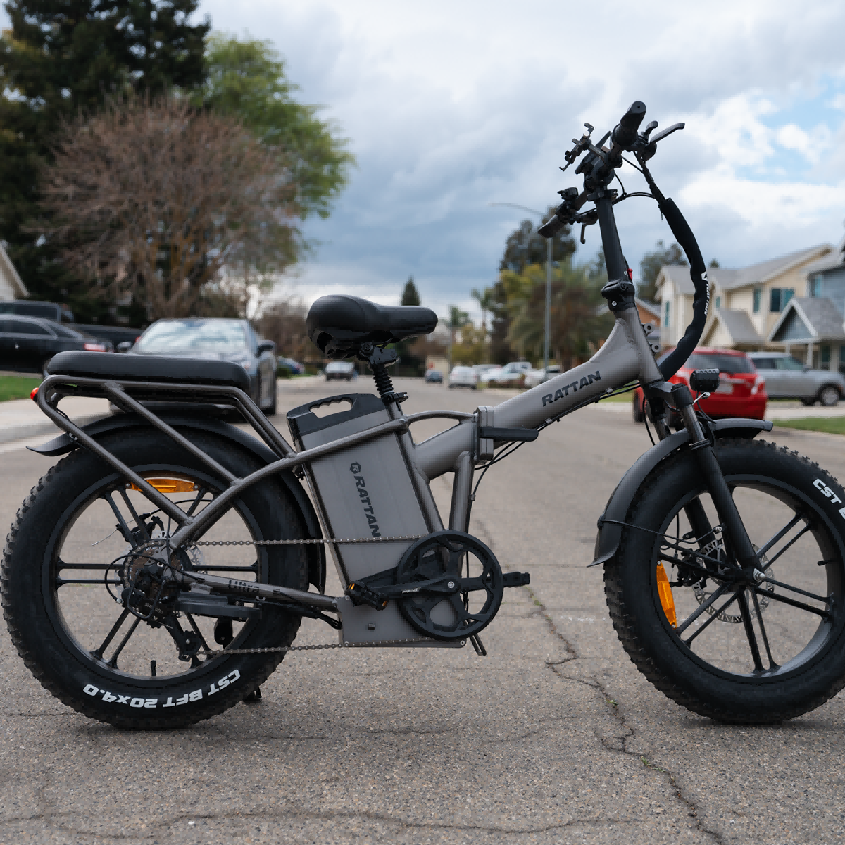 Szary rower elektryczny z szerokimi oponami zaparkowany na ulicy, domy w tle.