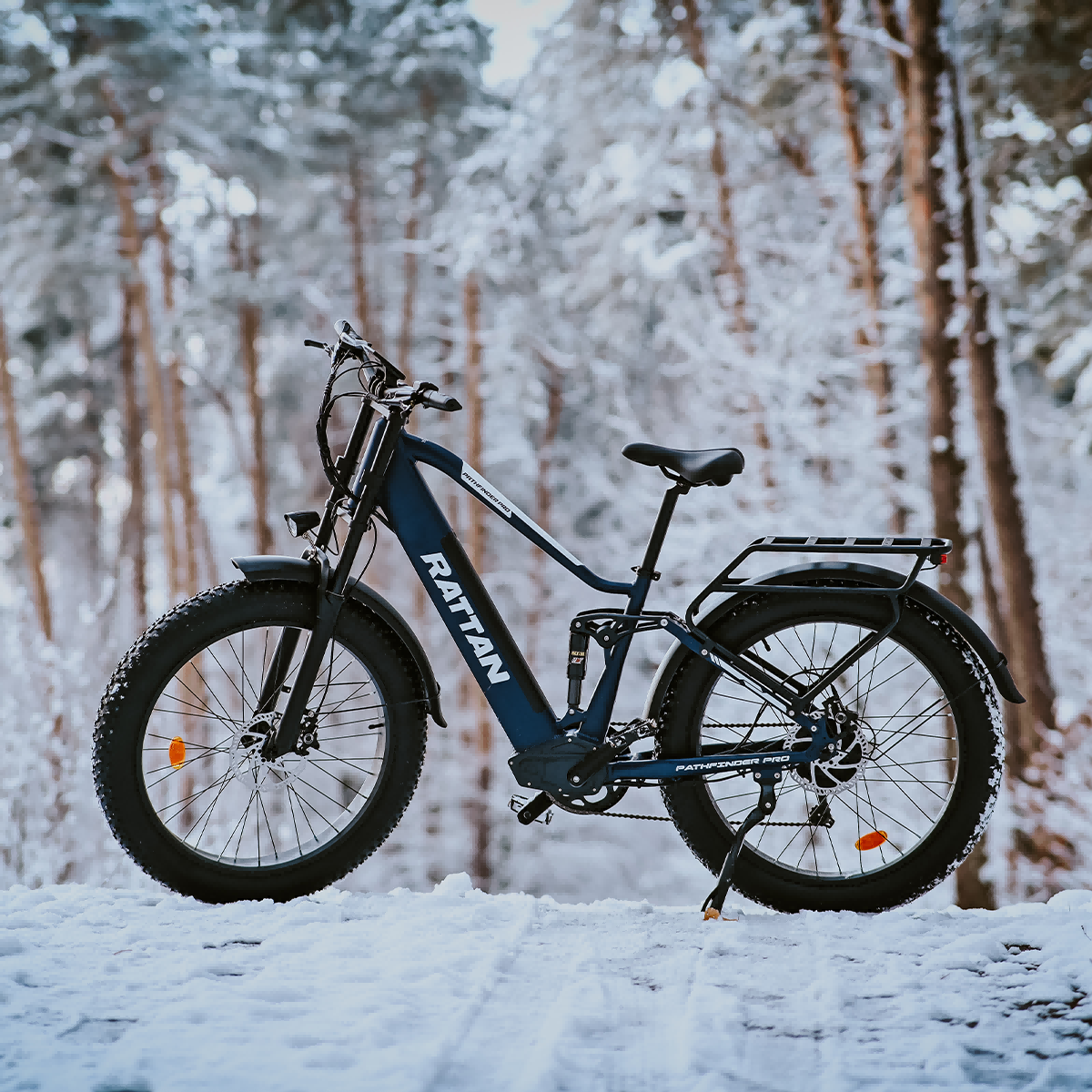 Ciemnoniebieski rower elektryczny z grubymi oponami stoi na śniegu przed zaśnieżonym lasem.
