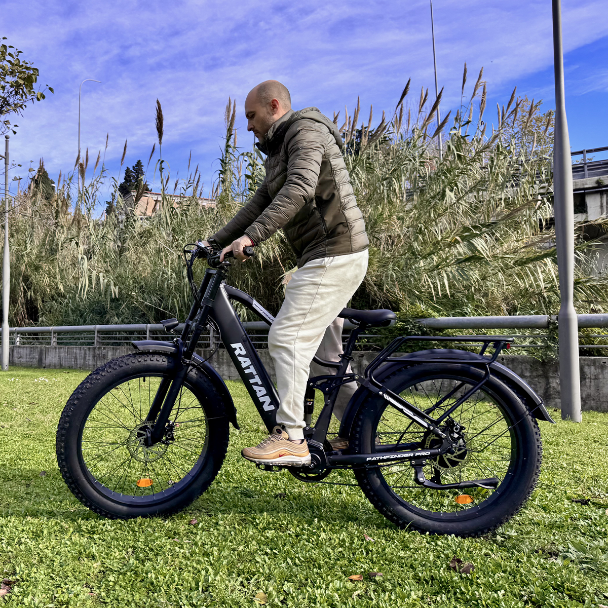 Mężczyzna jedzie na czarnym rowerze elektrycznym po trawniku, w tle trzciny i niebieskie niebo.