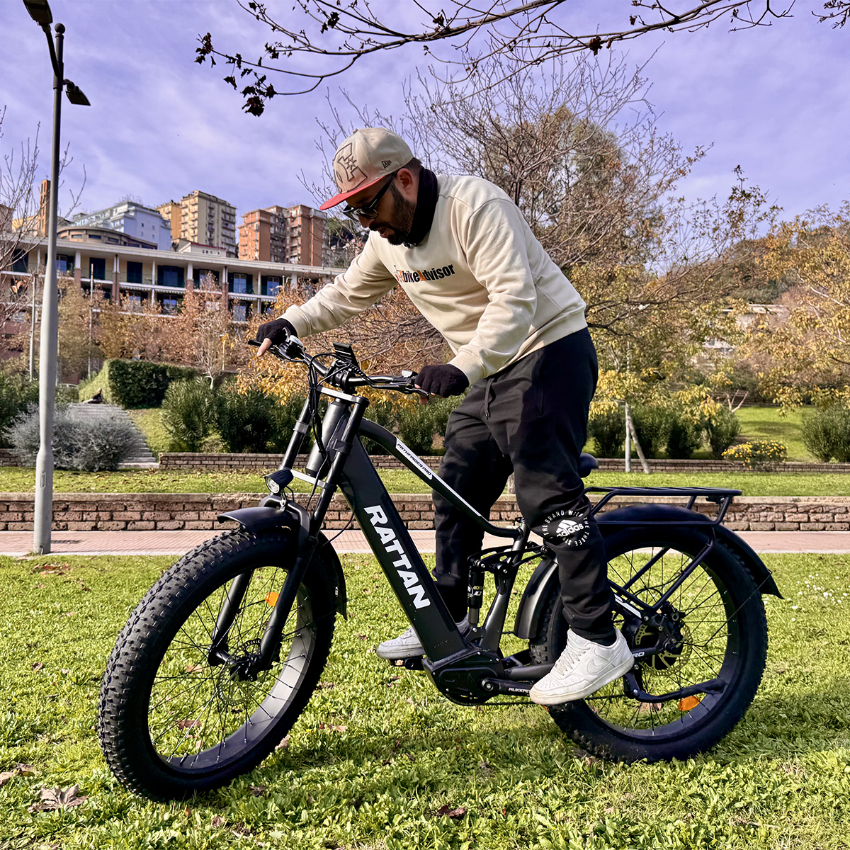 Mężczyzna w czapce i swetrze jedzie czarnym rowerem elektrycznym RATTAN po trawie. Budynki w tle.