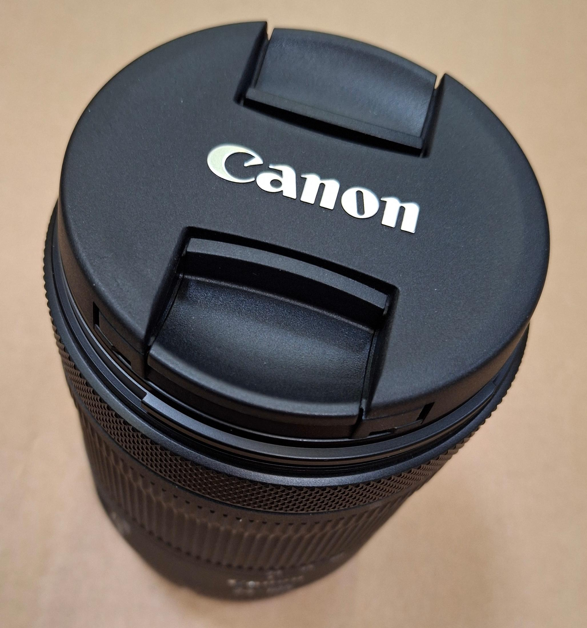 Tappo obiettivo Canon nero con logo argento su superficie marrone.