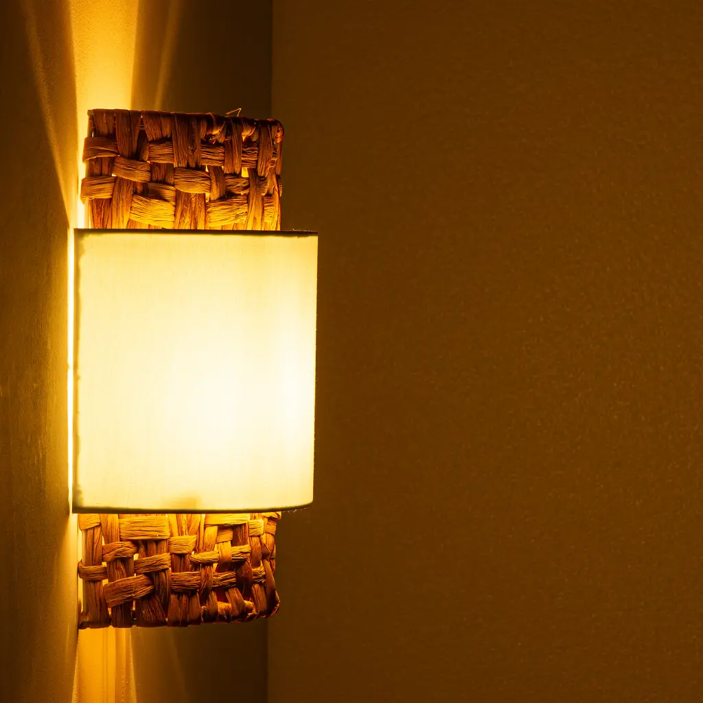 Una lámpara de pared con pantalla blanca y base de cesta tejida brilla amarilla.