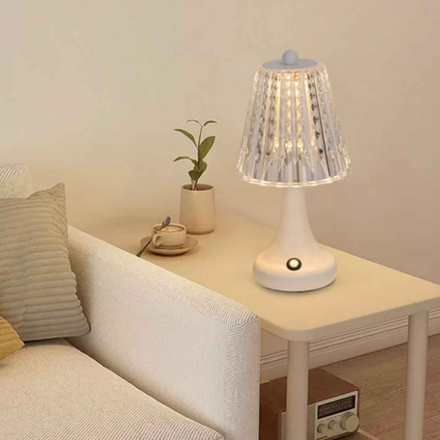 Eine weiße Lampe mit einem Kristalllampenschirm auf einem Beistelltisch neben einem Sofa.