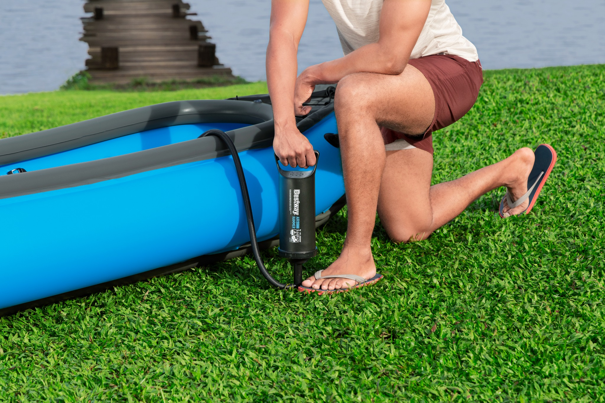 Person pumpt blauen Kajak auf Gras, See und Steg im Hintergrund.