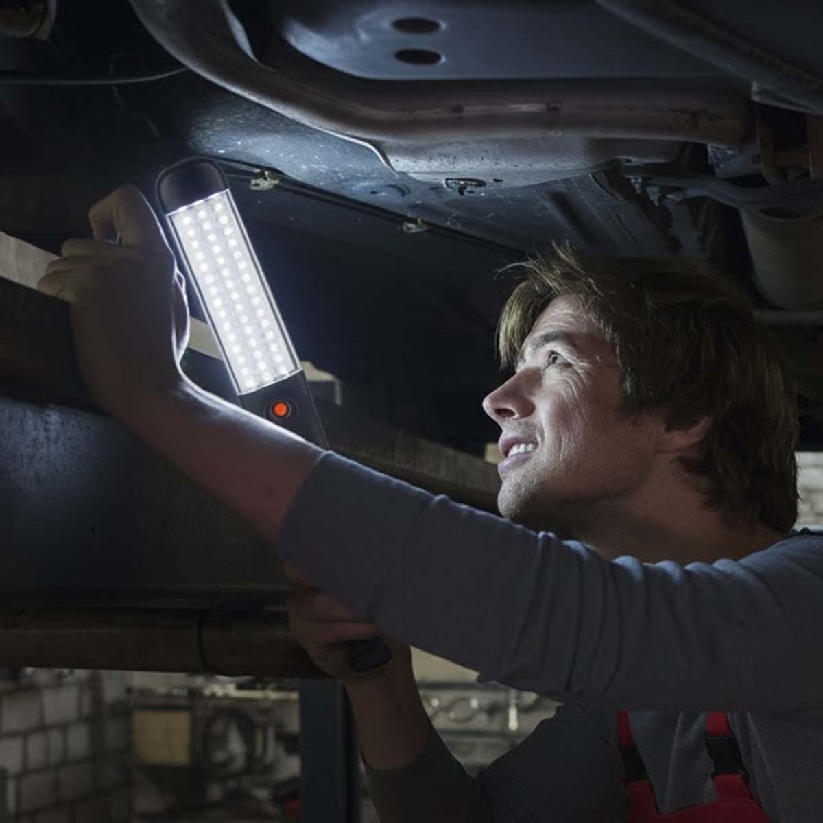 Ein Mann leuchtet mit einer Lampe unter ein Auto. Er trägt Arbeitskleidung.