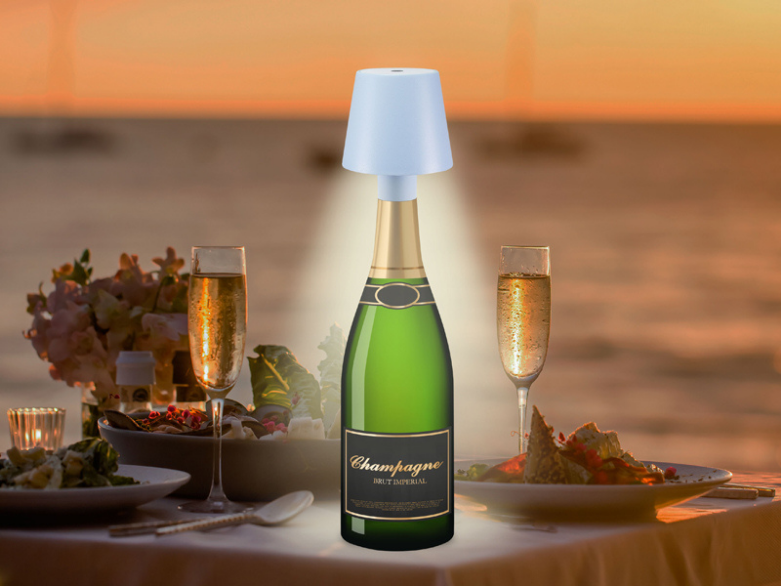 Bottiglia di champagne con lampada, bicchieri e cibo sulla spiaggia al tramonto.