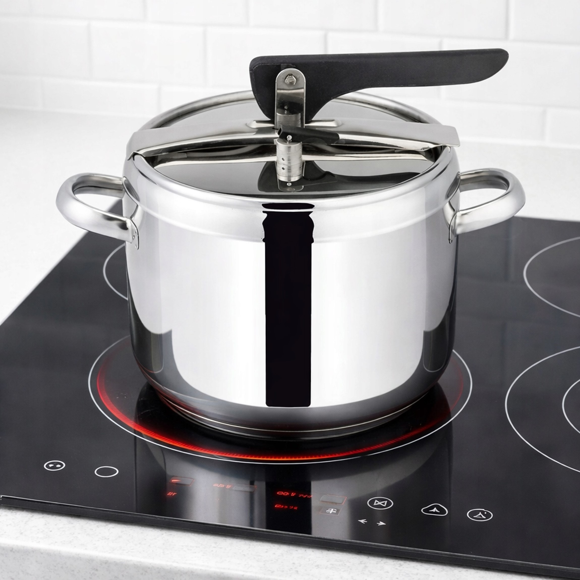 Błyszczący szybkowar ze stali nierdzewnej stoi na czarnej płycie indukcyjnej z czerwonymi elementami, na tle białych płytek.