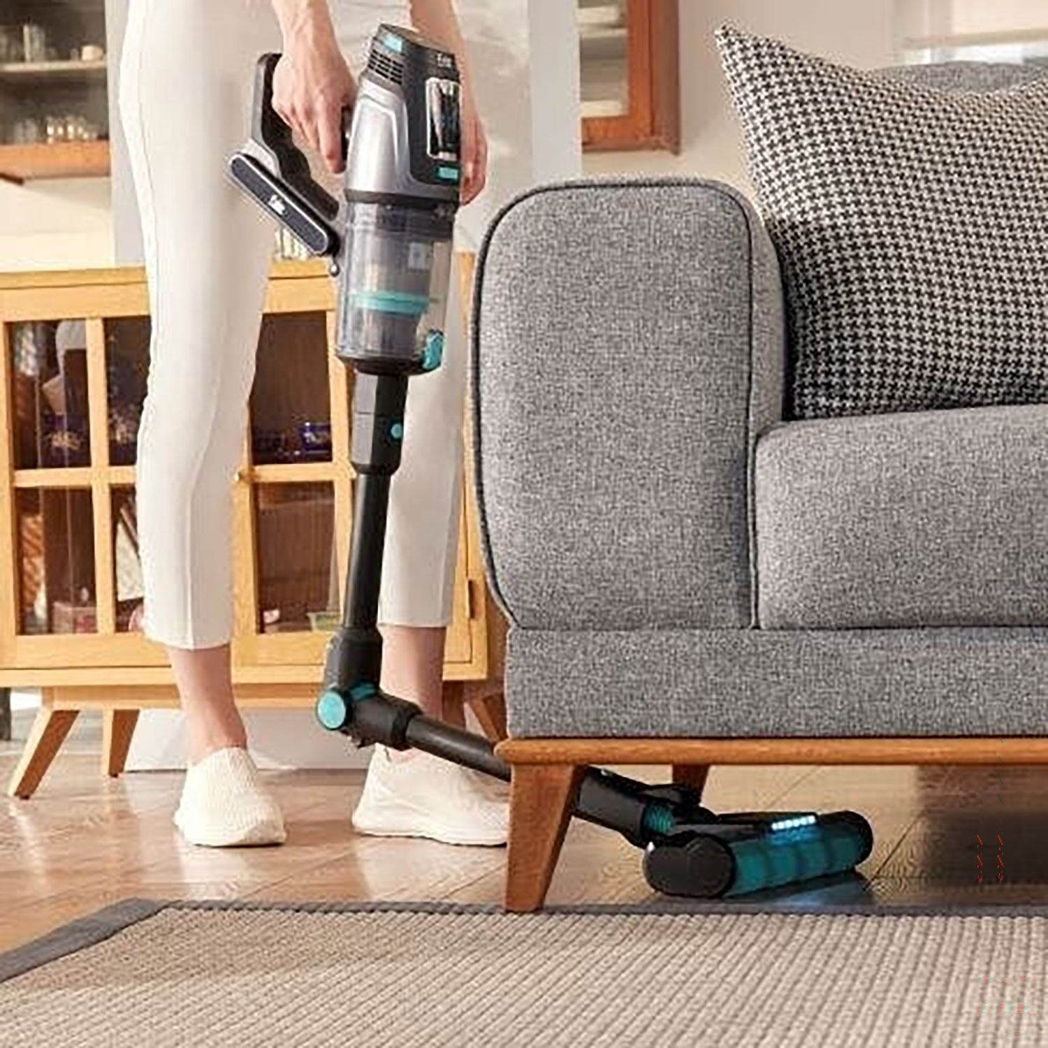 Eine Person benutzt einen Akku-Staubsauger, um unter einem grauen Sofa mit gemustertem Kissen zu reinigen.