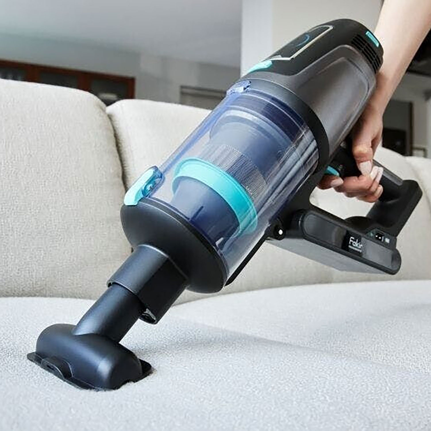 Eine Hand hält einen schwarzen Akku-Staubsauger, der auf einem hellen Sofa verwendet wird.