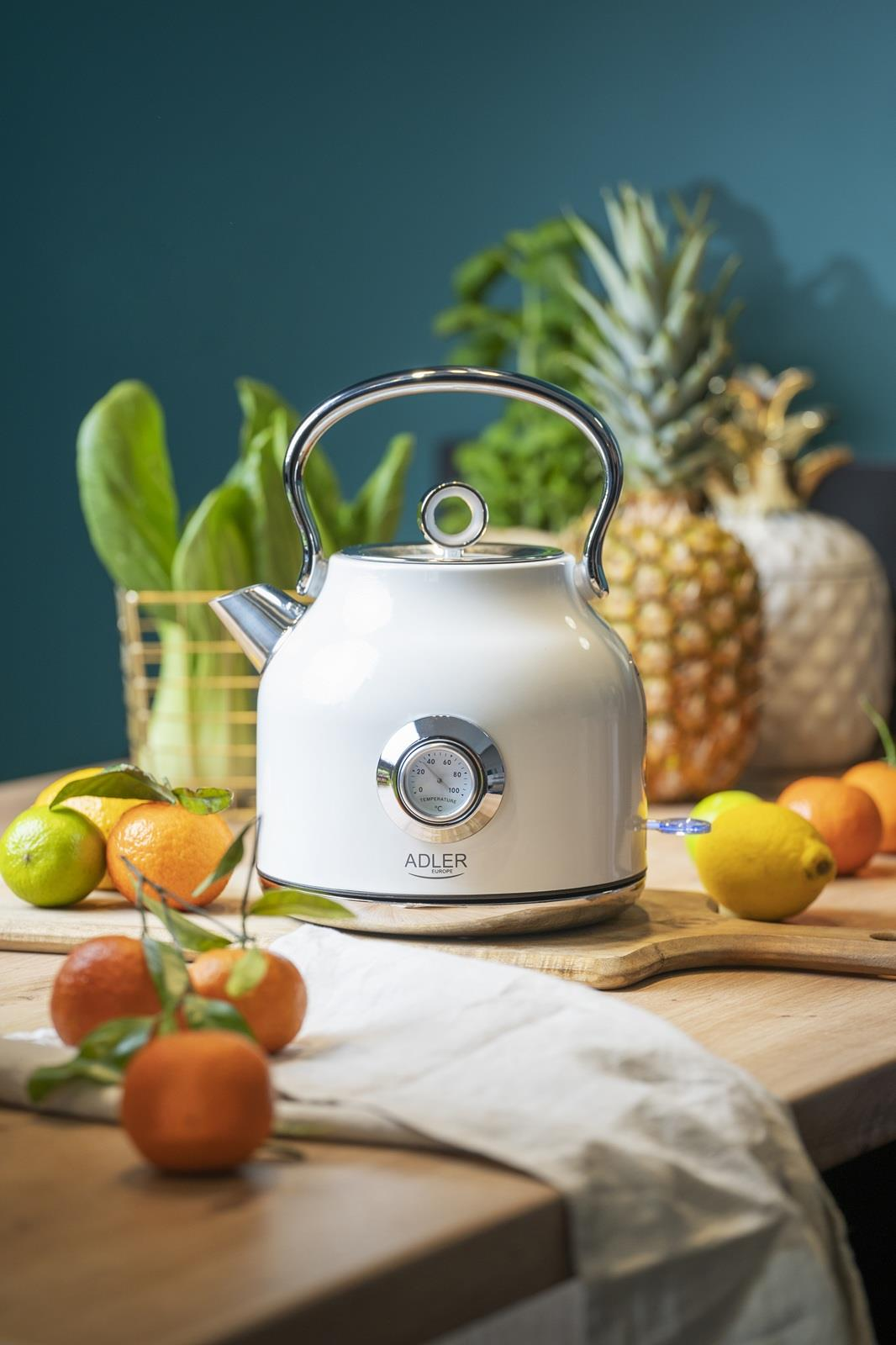 Biały czajnik z termometrem stoi na drewnianym stole obok owoców.