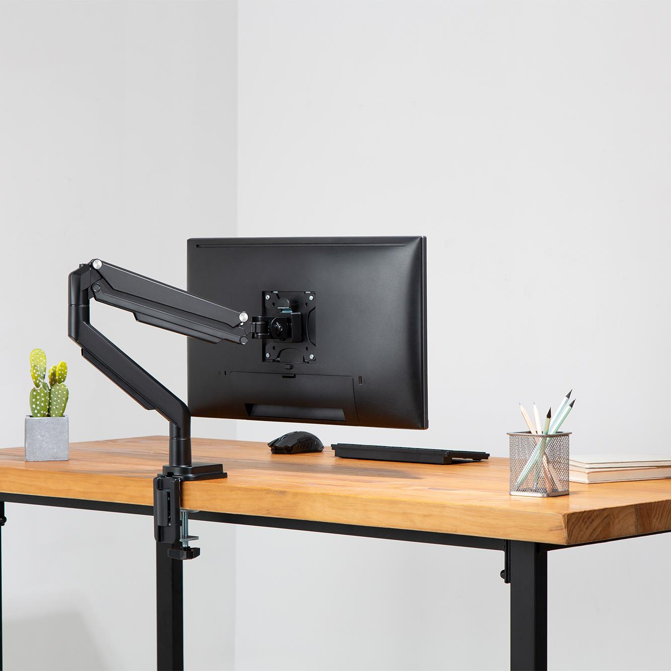 Czarny monitor zamontowany na biurku za pomocą ramienia. Widoczny kaktus i mysz.